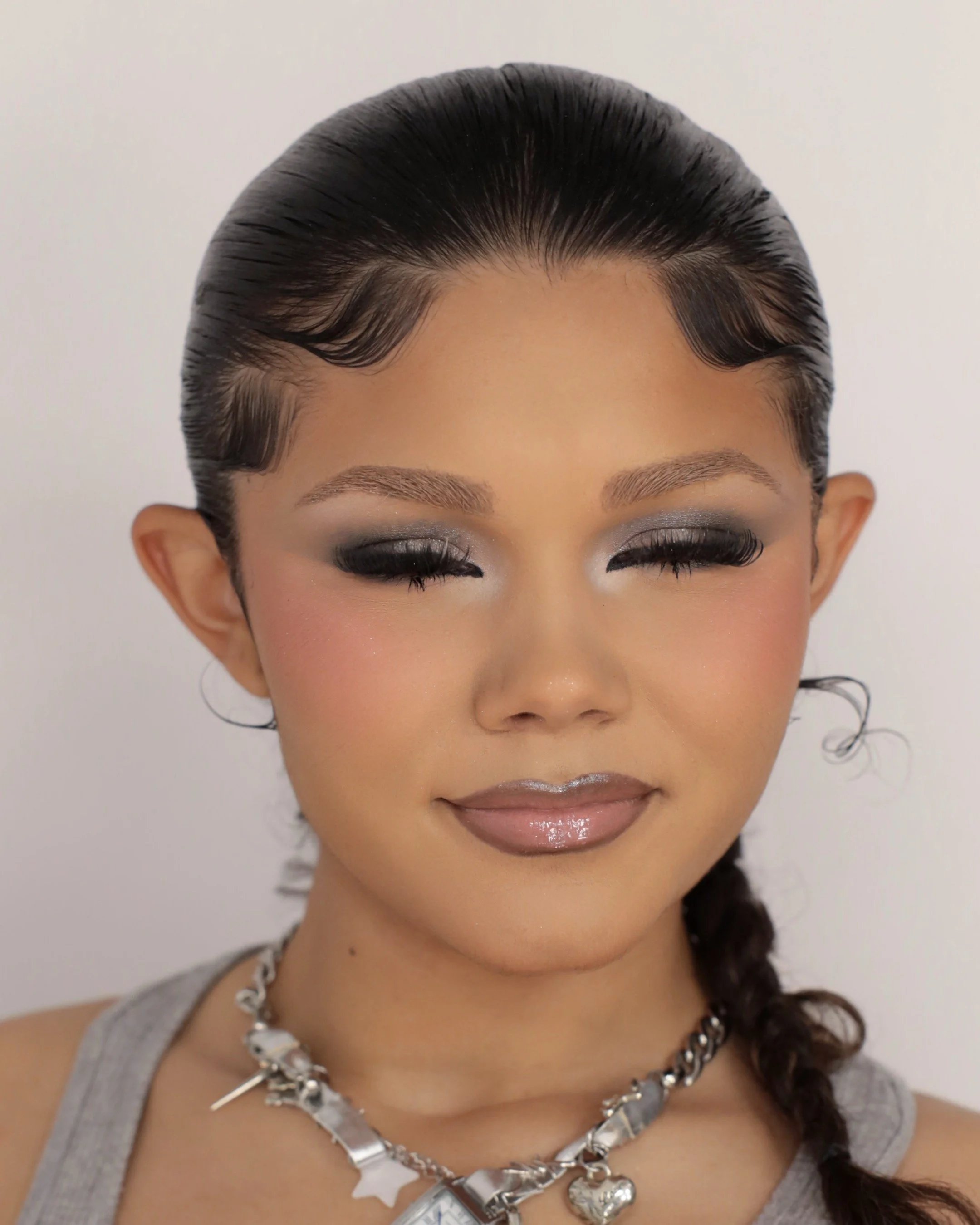 Close-up of a woman's face with styled hair, makeup, and layered silver necklaces, wearing a gray sleeveless top.