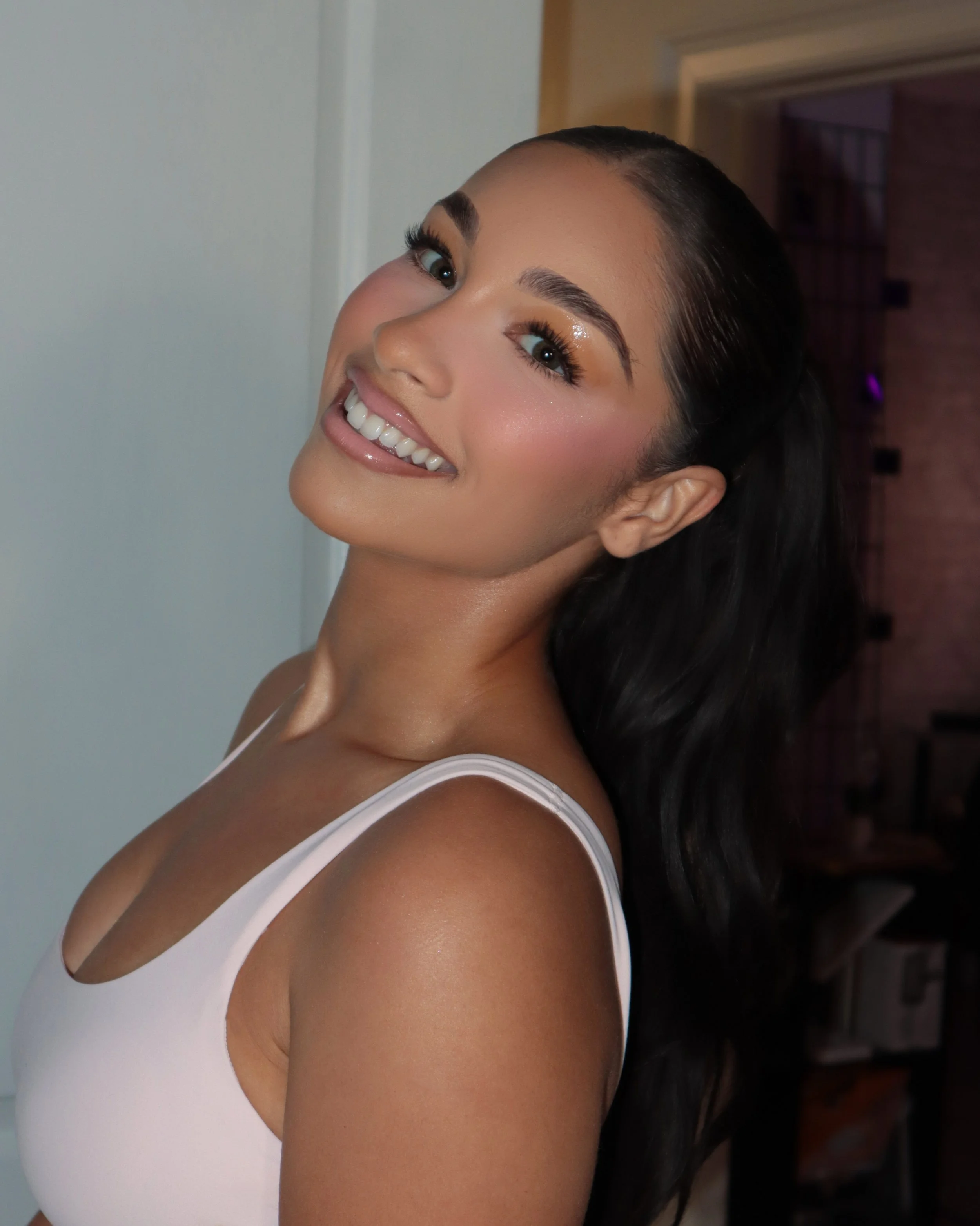 Close-up of a smiling woman with dark hair, wearing a white sleeveless top, in front of a plain wall.