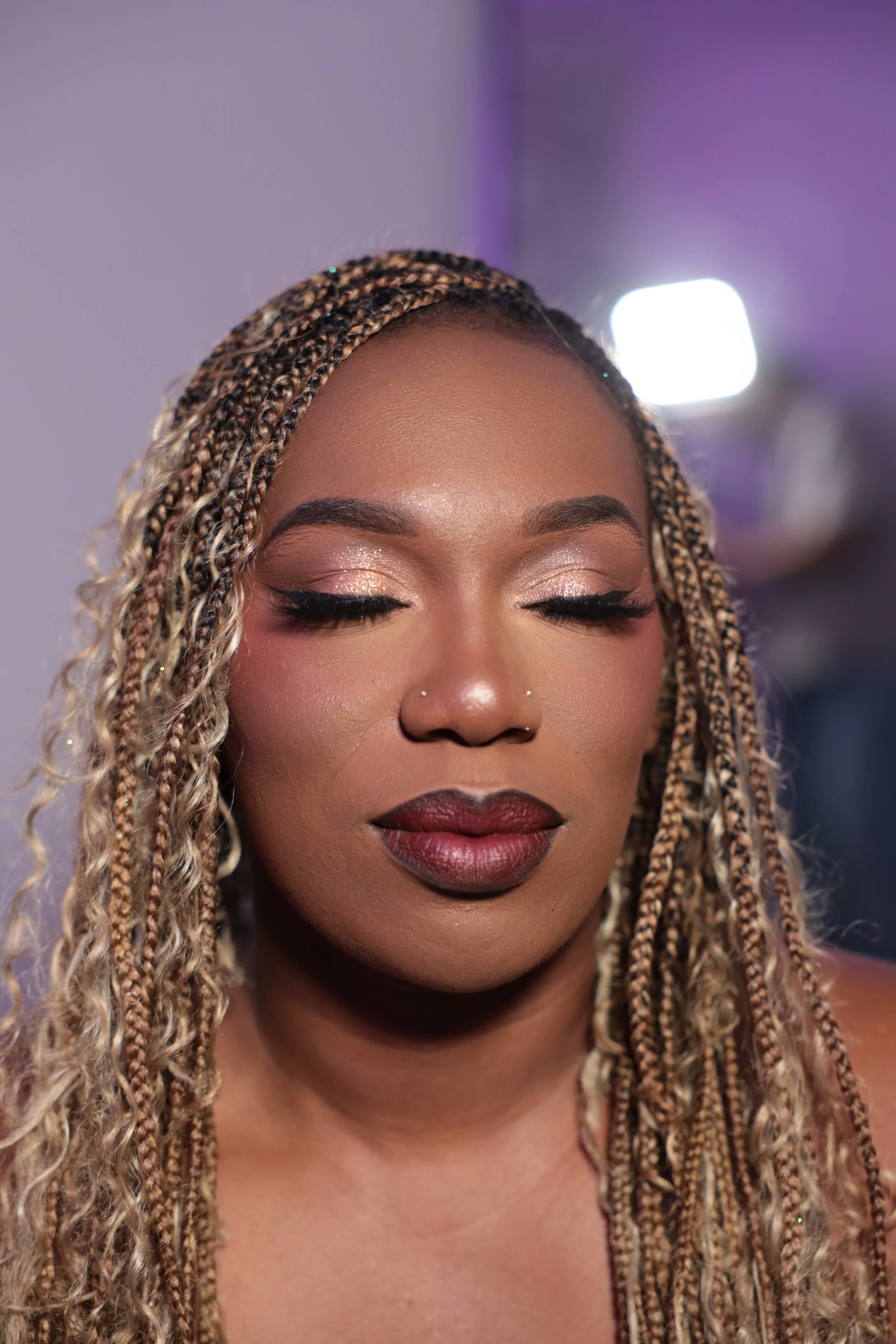 Close-up of a woman with braided hair and makeup with her eyes closed, showing shimmering eyeshadow and dark lipstick. The background has a purple hue with bright lights.