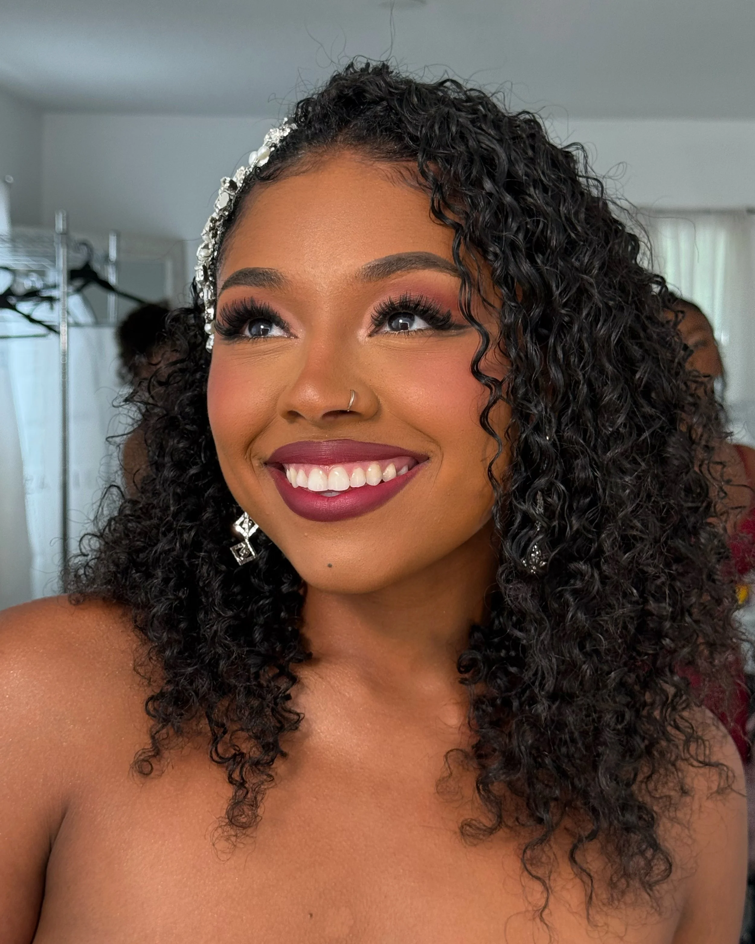 Close-up of a smiling woman with curly hair, wearing earrings and makeup, indoors with a blurred background.