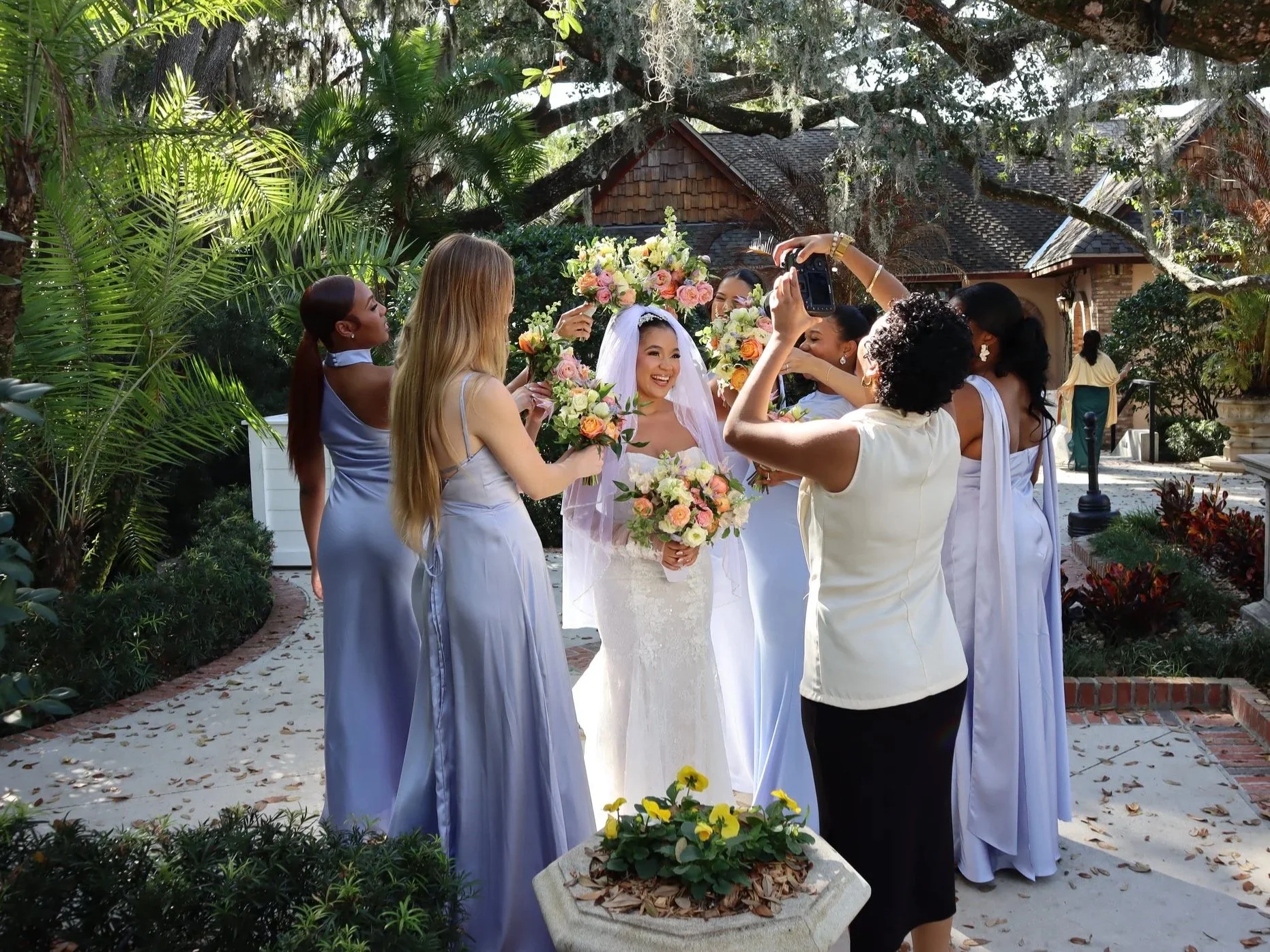 A bride in a white wedding dress and veil surrounded by bridesmaids in light purple dresses, holding bouquets of pink and white flowers, during a wedding celebration outdoors with greenery and a house in the background.