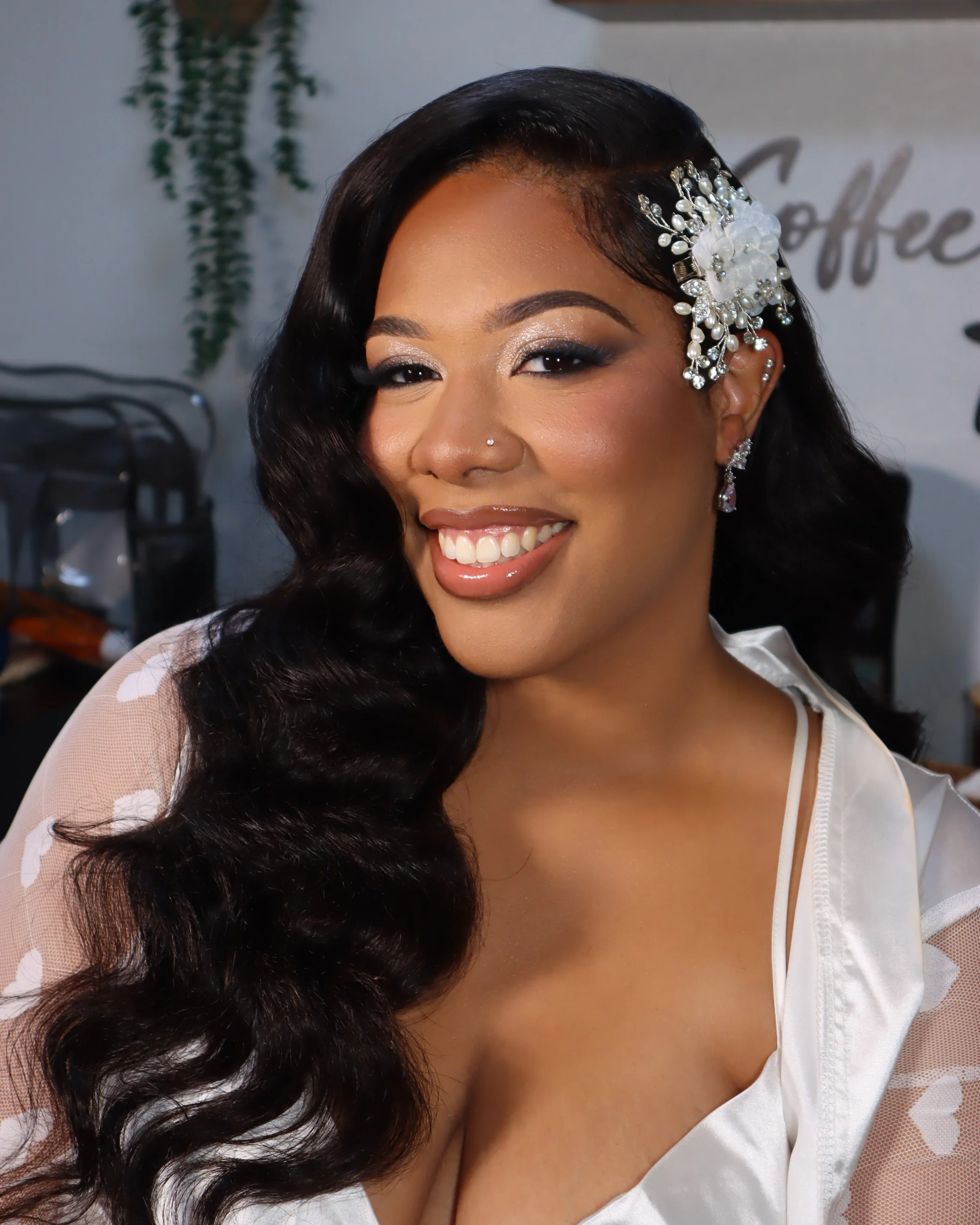 A smiling woman with long, wavy dark hair adorned with a decorative white floral hairpiece with pearls and rhinestones, wearing makeup including shimmery eyeshadow and lipstick, dressed in a white top with sheer sleeves and a low neckline.