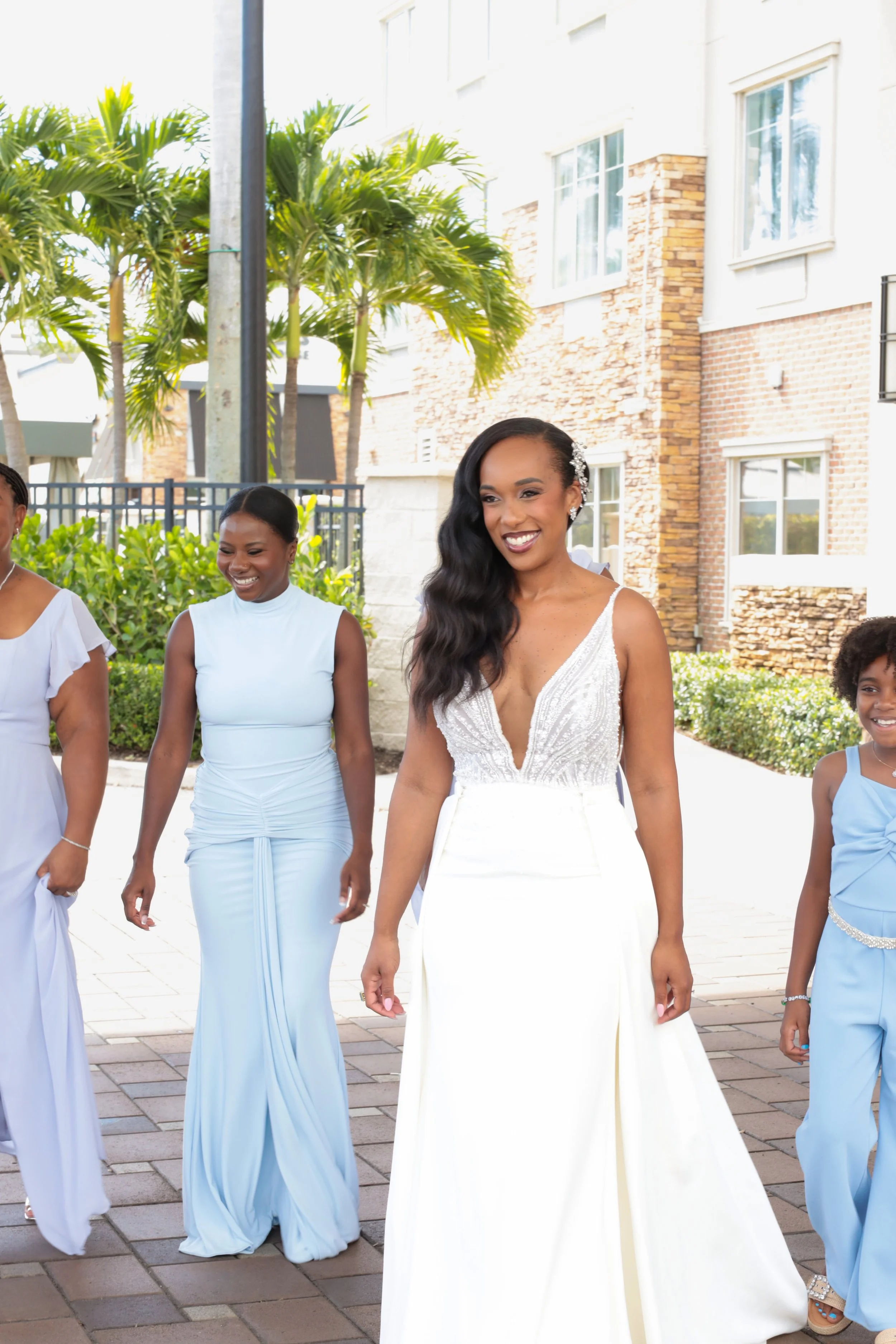 A group of women, one in a white wedding dress with long black wavy hair and floral hair accessory, surrounded by women in light blue dresses, walking outdoors on a paved path with palm trees and modern brick buildings in the background.