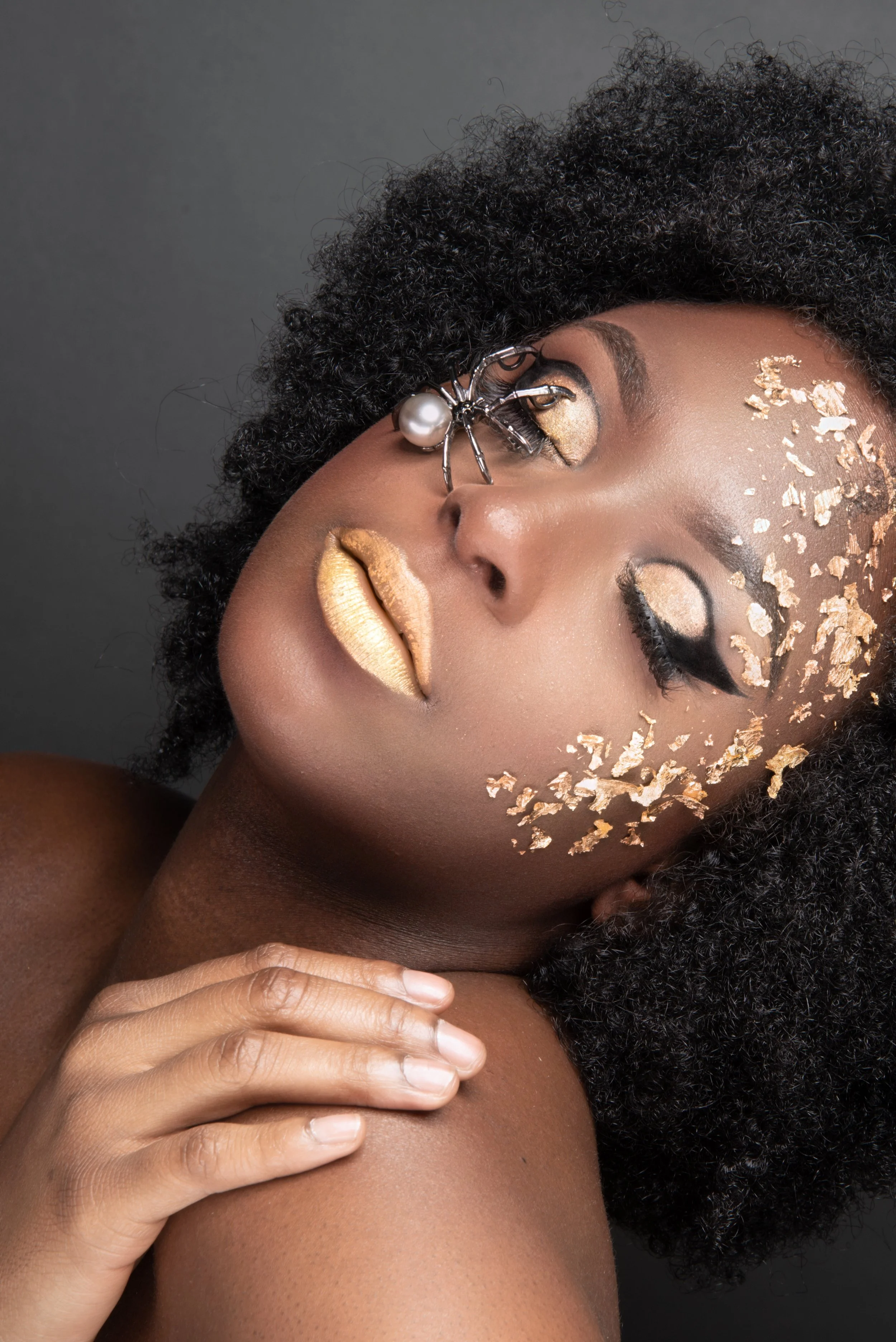 A close-up of a woman with dark skin and curly hair, adorned with gold makeup, gold-colored lips, and gold flakes on her face, her eyes partially closed with gold eyeshadow.