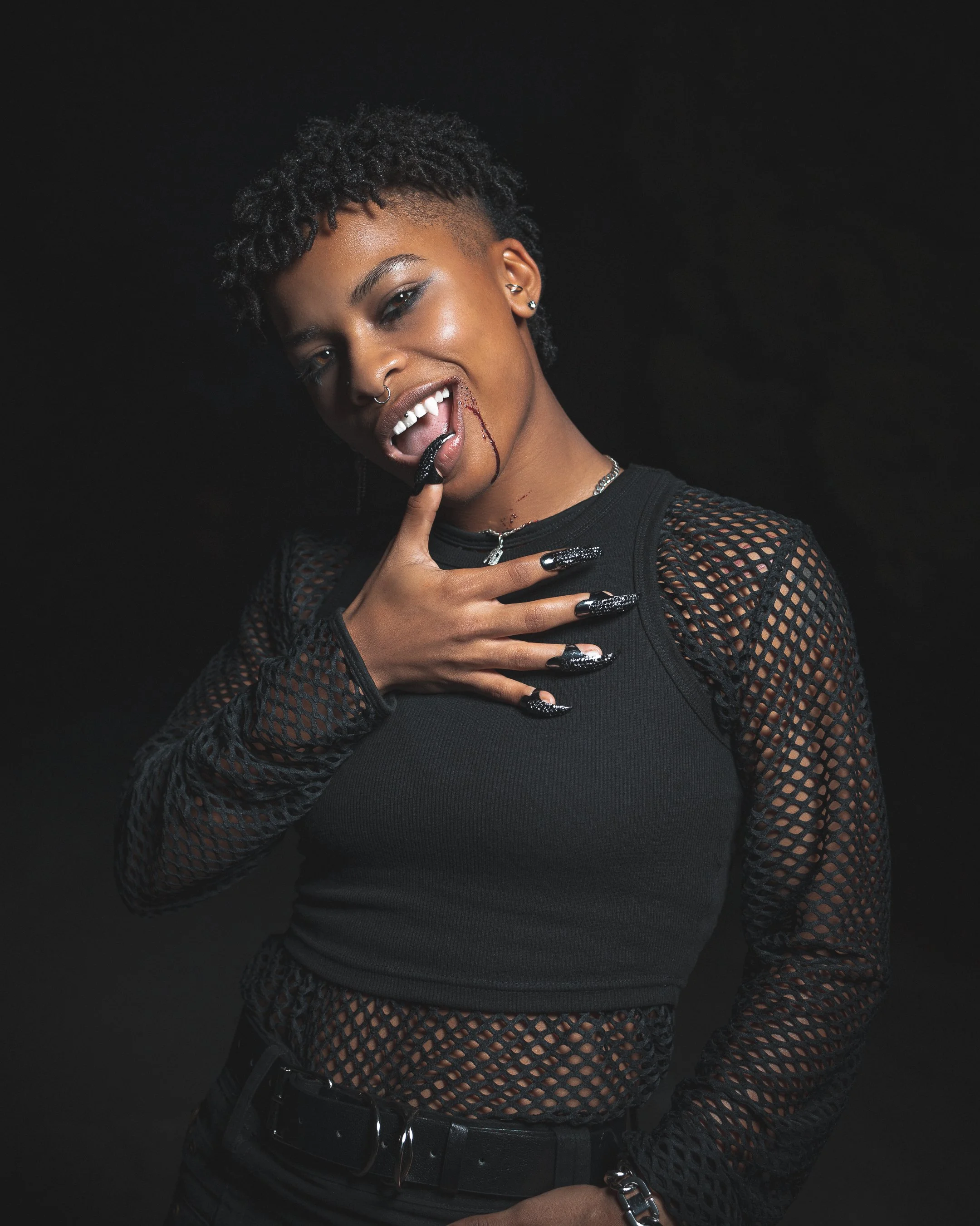 A woman with short, textured black hair, multiple earrings, and a septum piercing, is posing against a black background. She wears a black mesh top over a black shirt, black pants, and a belt, and is holding a black whip close to her mouth, with bloo