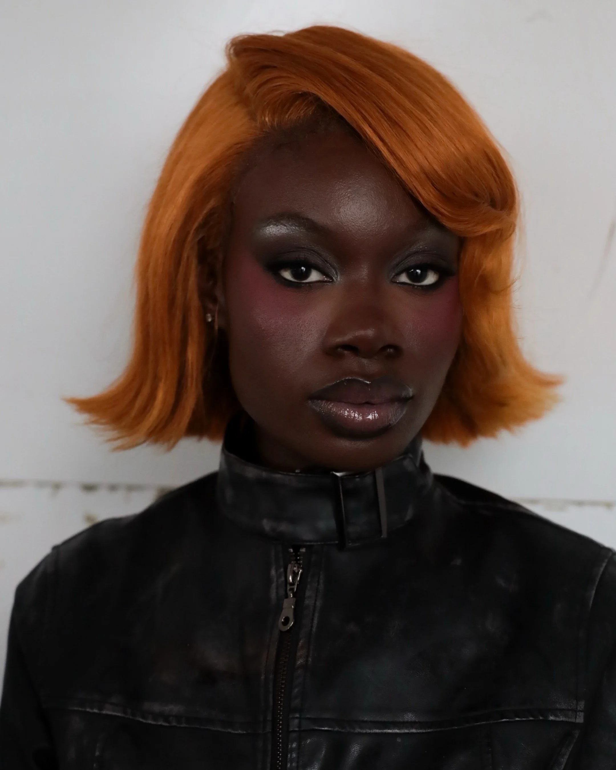 Close-up of a woman with dark skin, orange hair styled in a bob, wearing a black leather jacket, against a plain neutral background.