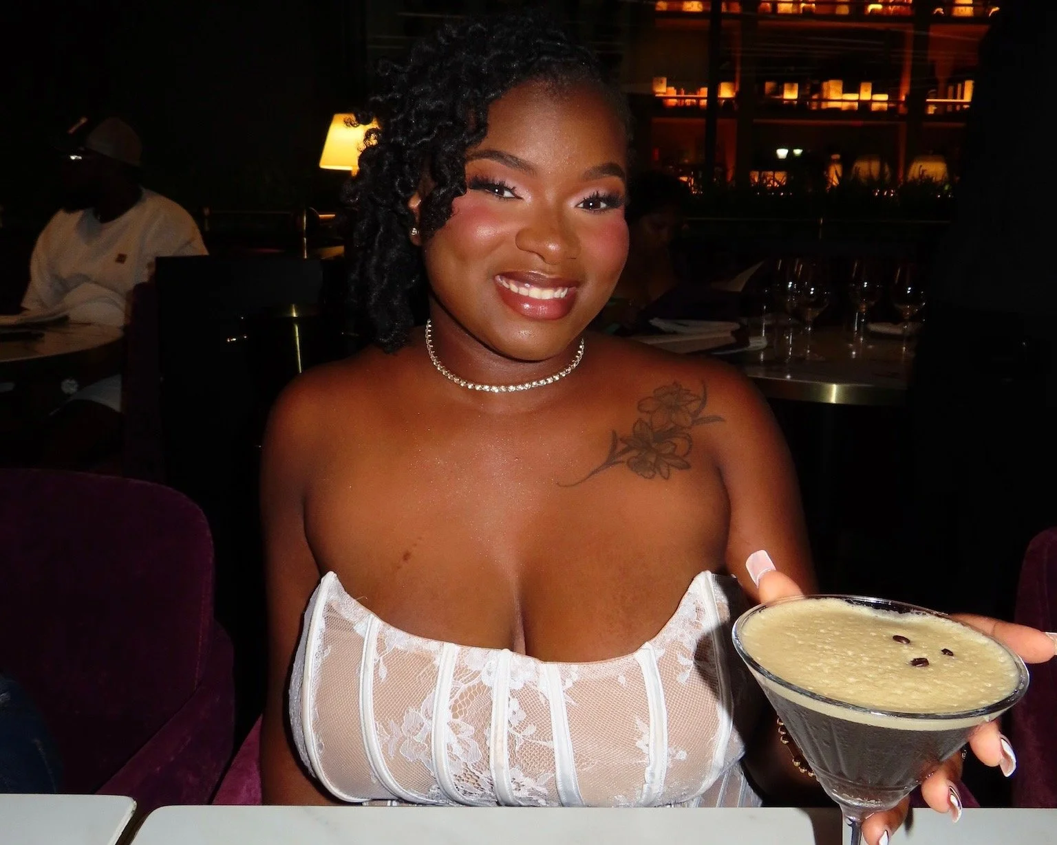 A woman with curly hair, wearing a pearl choker and a white strapless top, is smiling and holding a cocktail in a martini glass at a restaurant.