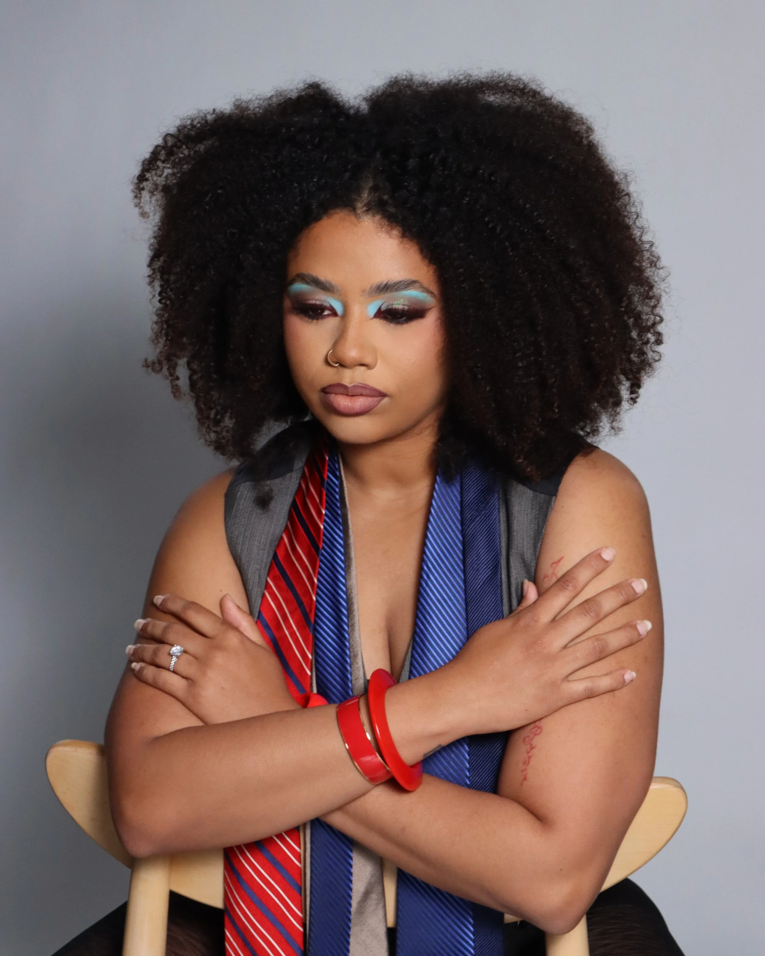 A woman with curly hair and makeup with bright blue eyeshadow, sitting with her arms crossed on a chair, wearing red jewelry and a metallic top, against a plain gray background.