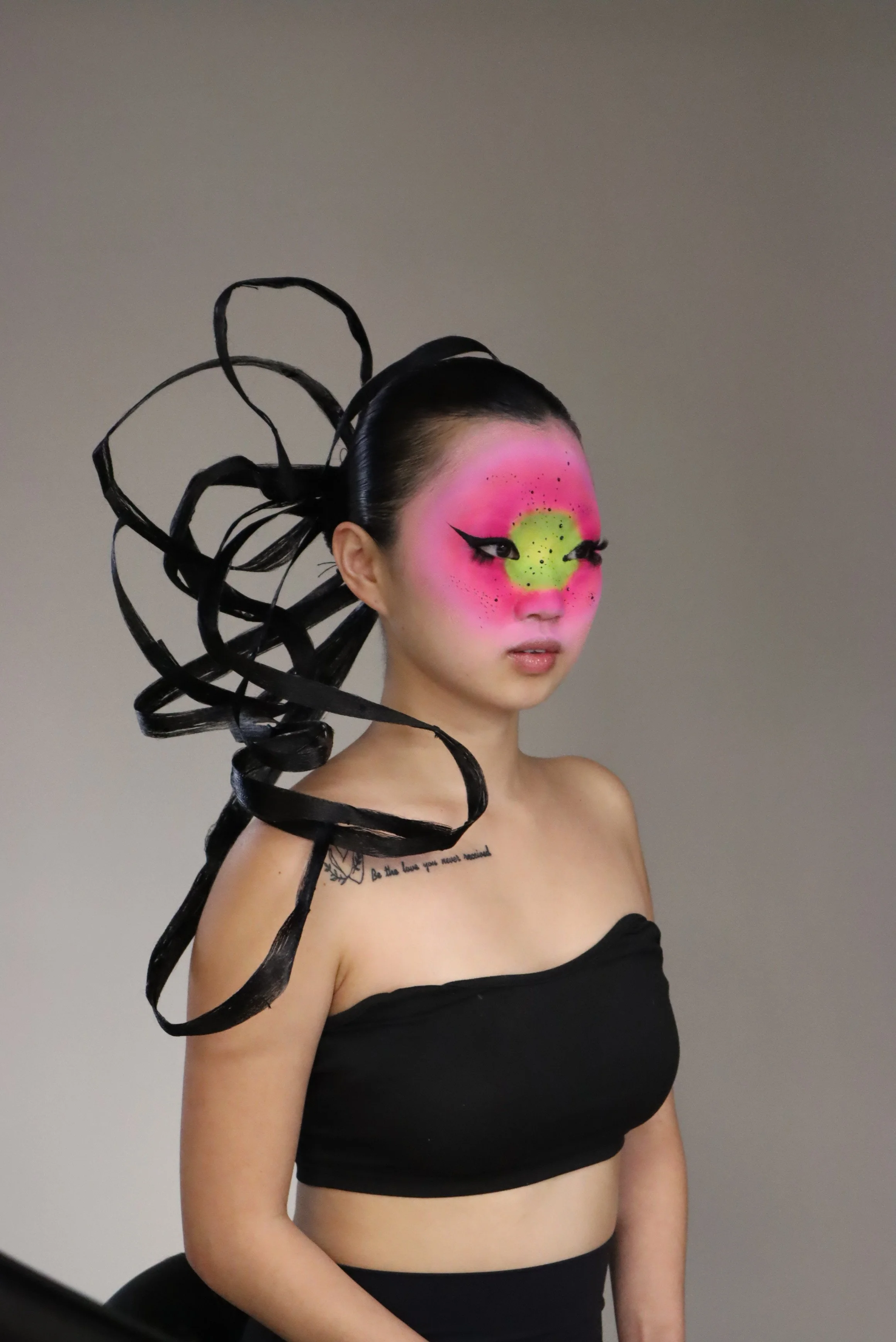 A woman with artistic makeup resembling a pink and green alien face, black dramatic eyeliner, and black ribbon hair accessories, wearing a black strapless top.