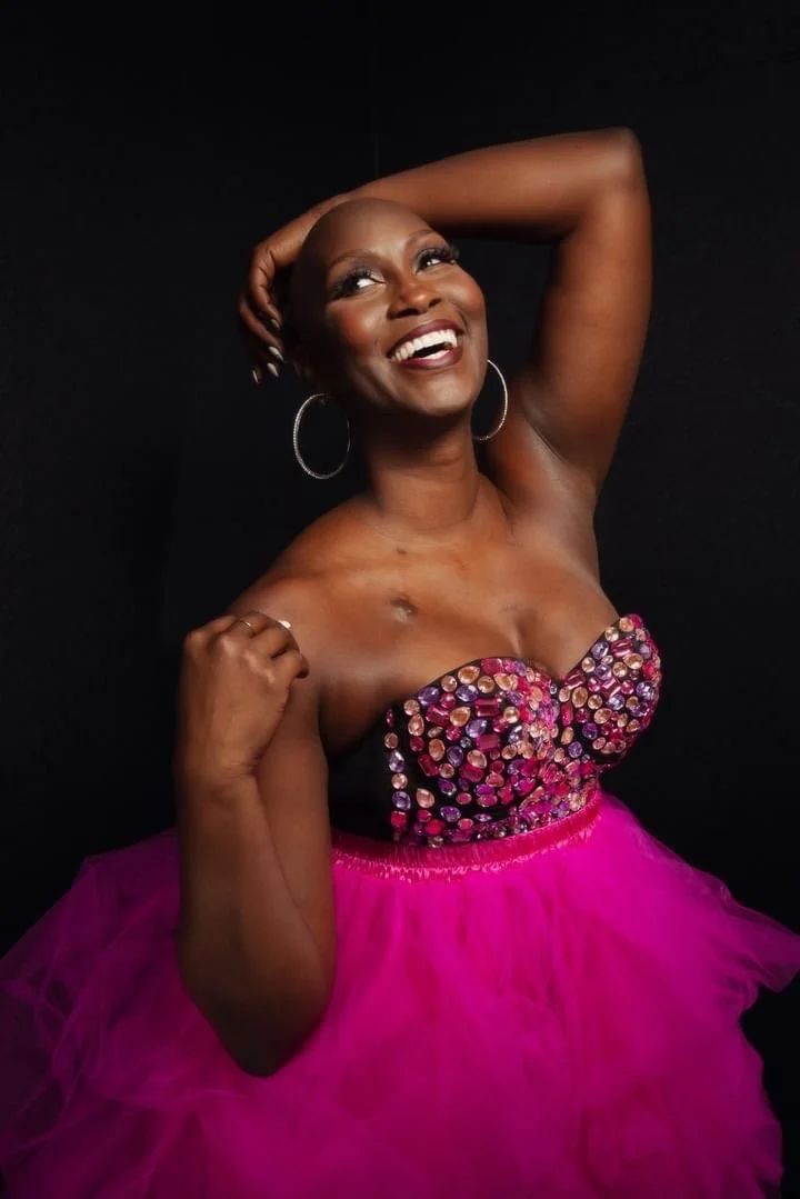 A woman with a bald head wearing large hoop earrings and a strapless dress with a bejeweled bodice and a pink tulle skirt, smiling and posing against a black background.