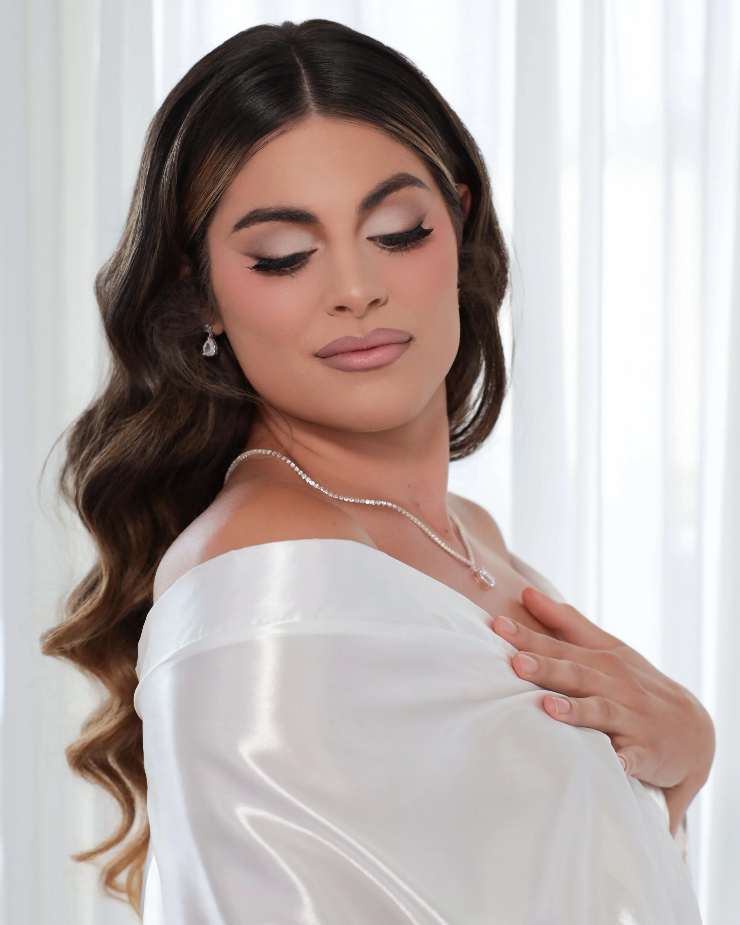 A woman with long, wavy brown hair, closed eyes, and wearing makeup, is dressed in white satin, with a pearl necklace, standing in front of white curtains.