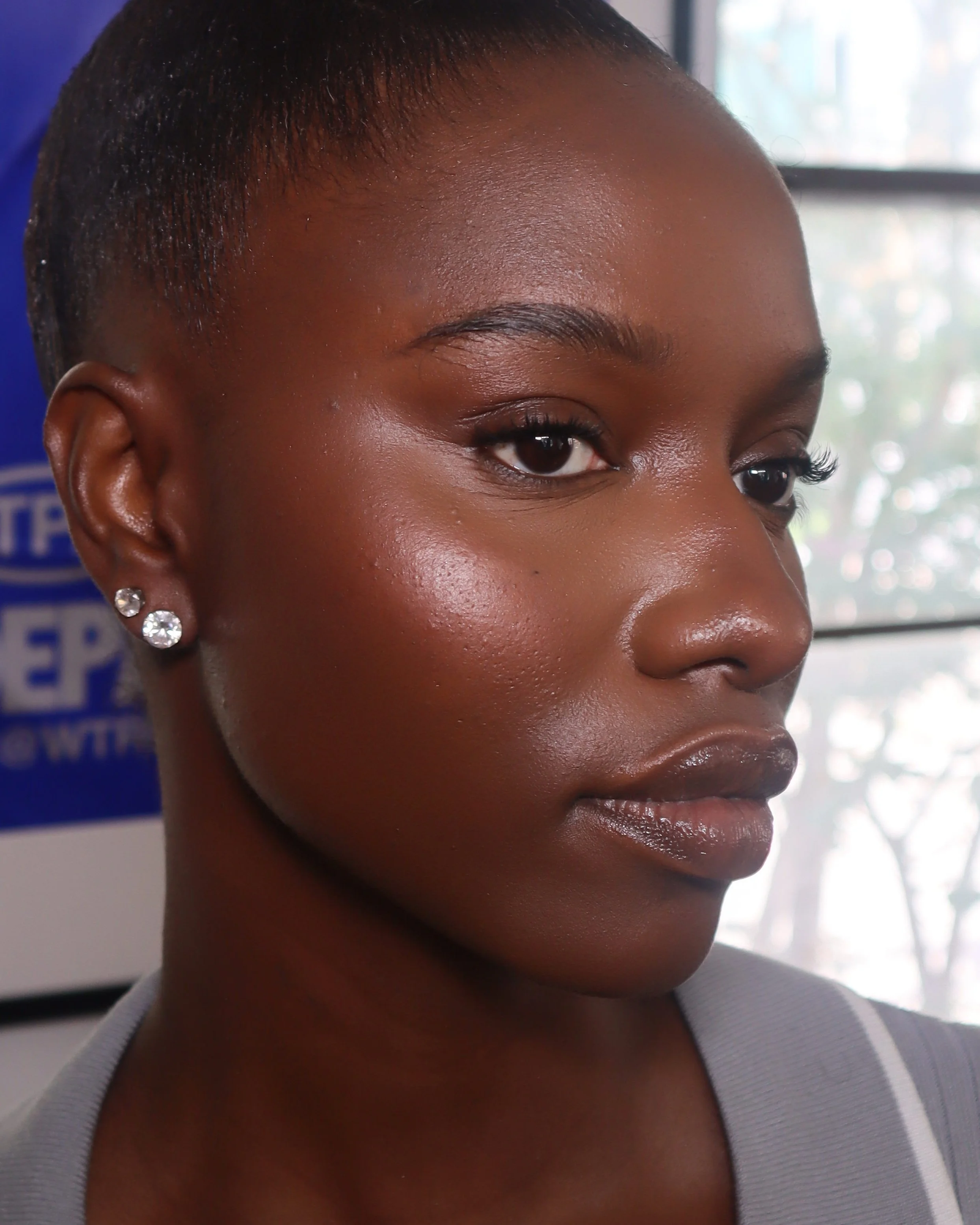 Close-up of a woman with short, shaved hairstyle, dark skin, and wearing diamond earrings, in an indoor setting with natural light coming through a window.