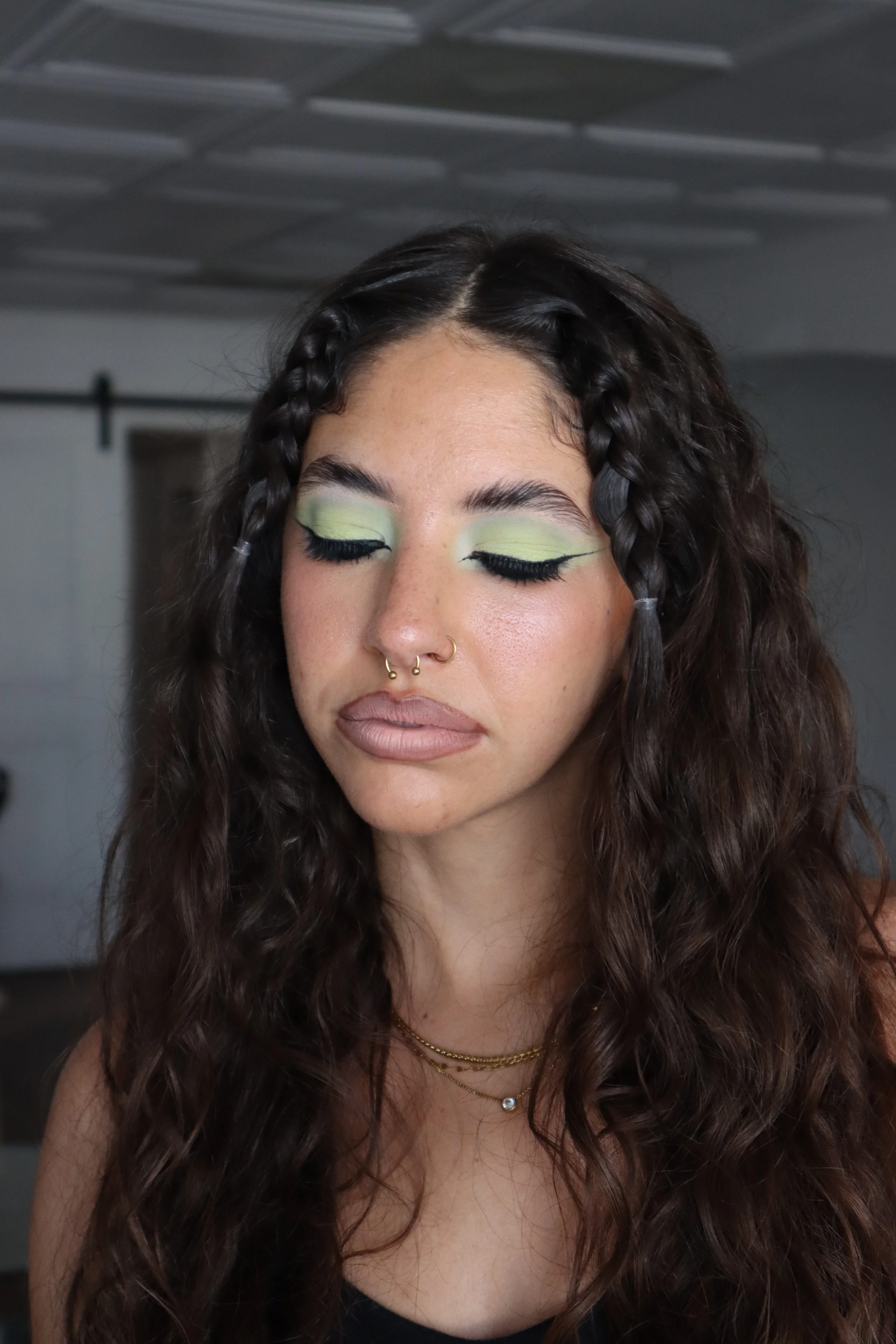 A young woman with long, curly brown hair and braided front sections, wearing makeup with green eyeshadow and black eyeliner, has her eyes closed. She is also wearing gold layered necklaces and has a septum piercing, standing in a room with a dimly l