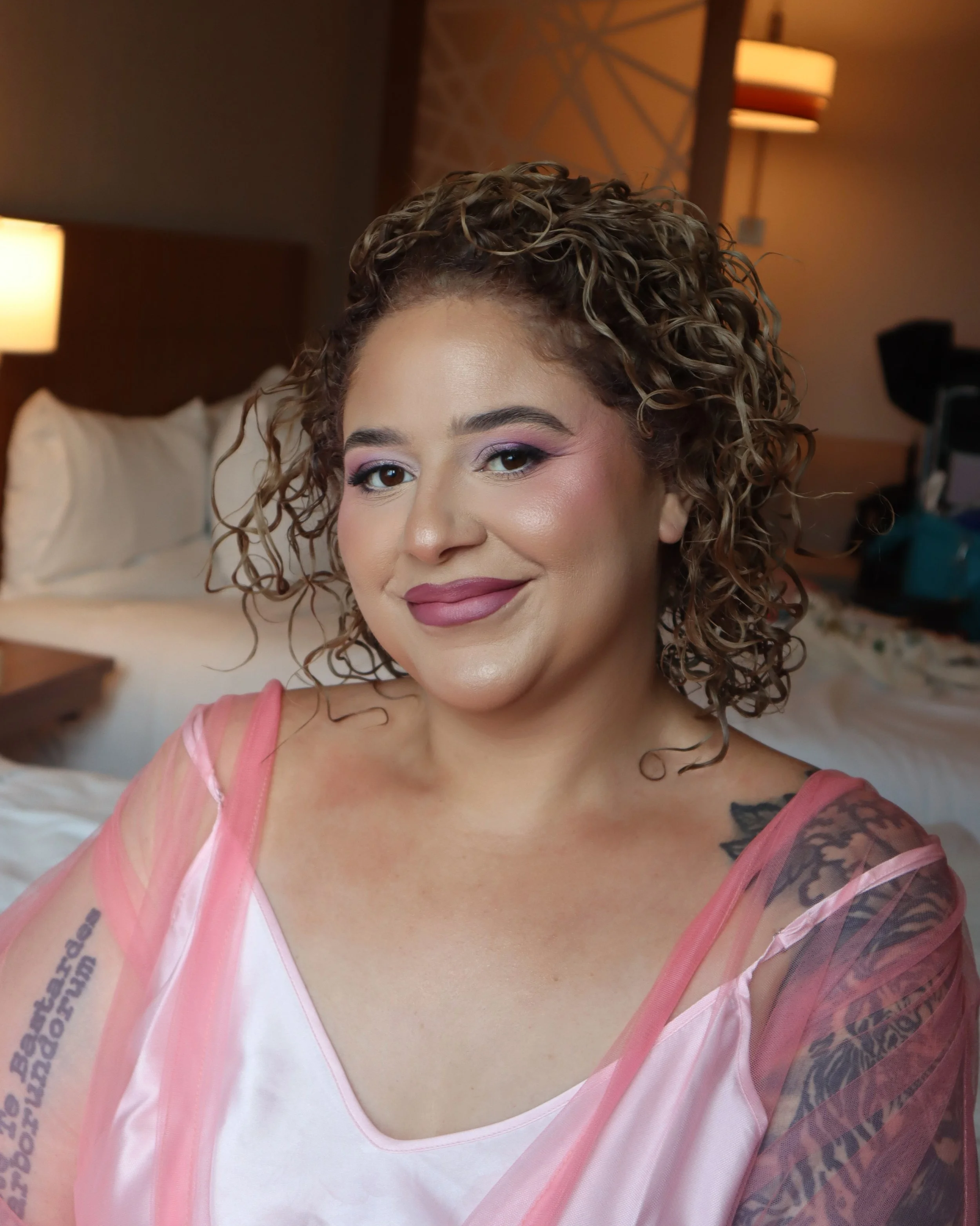 A woman with curly hair and tattoos, wearing a pink sheer top over a white shirt, smiling in a warmly lit hotel room.
