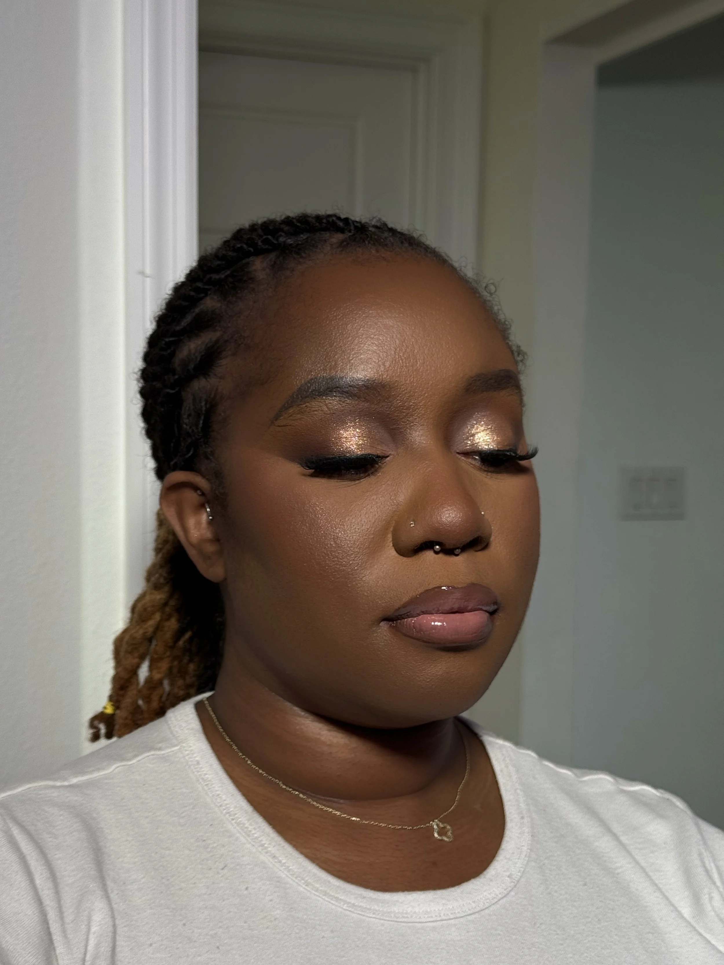 Close-up of a woman with braided hair, wearing makeup with gold eyeshadow, a nose piercing, a necklace with a heart pendant, and a white top, standing in a doorway.