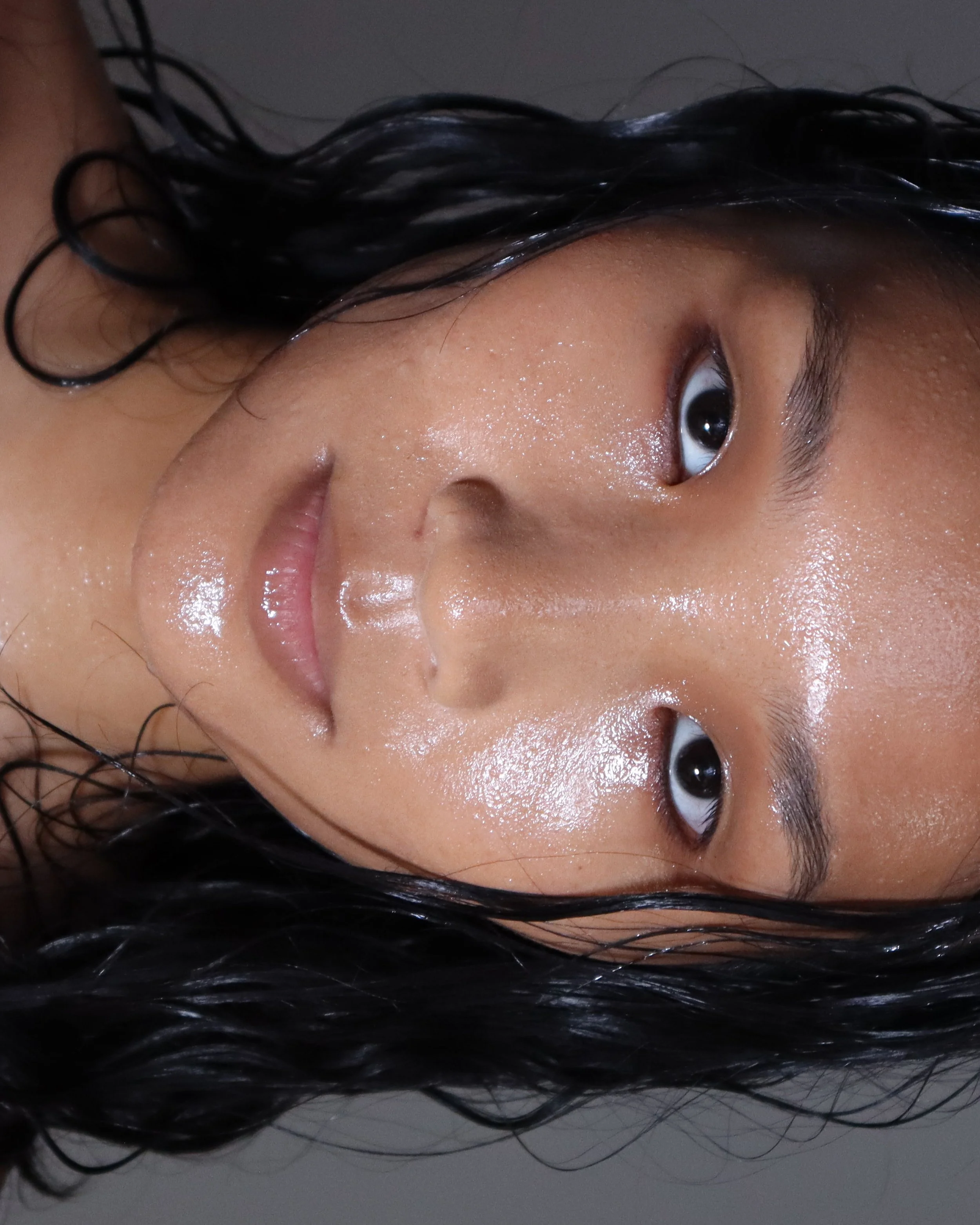 Close-up image of a young woman with wet, black, curly hair, looking directly at the camera with her finger touching her lips.
