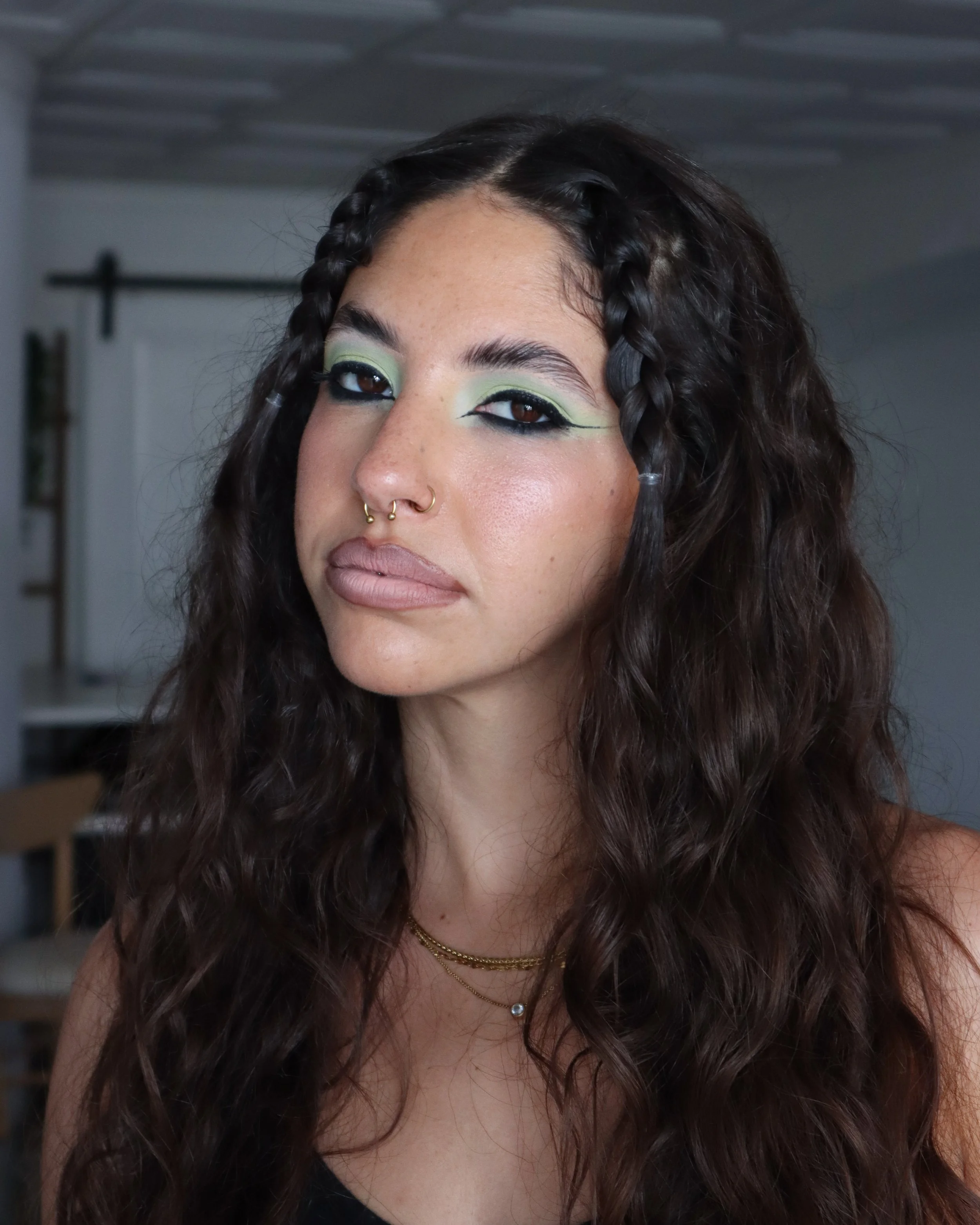 Close-up of a woman with dark curly hair, wearing neon green eye makeup, black eyeliner, and nude lipstick, accessorized with multiple gold necklaces and a nose ring.