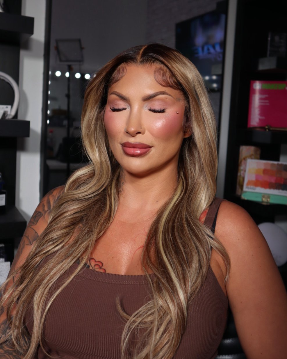 A woman with long, wavy blonde hair and light makeup, including glossy lips, with her eyes closed, in a room with shelves and a large window or mirror behind her.