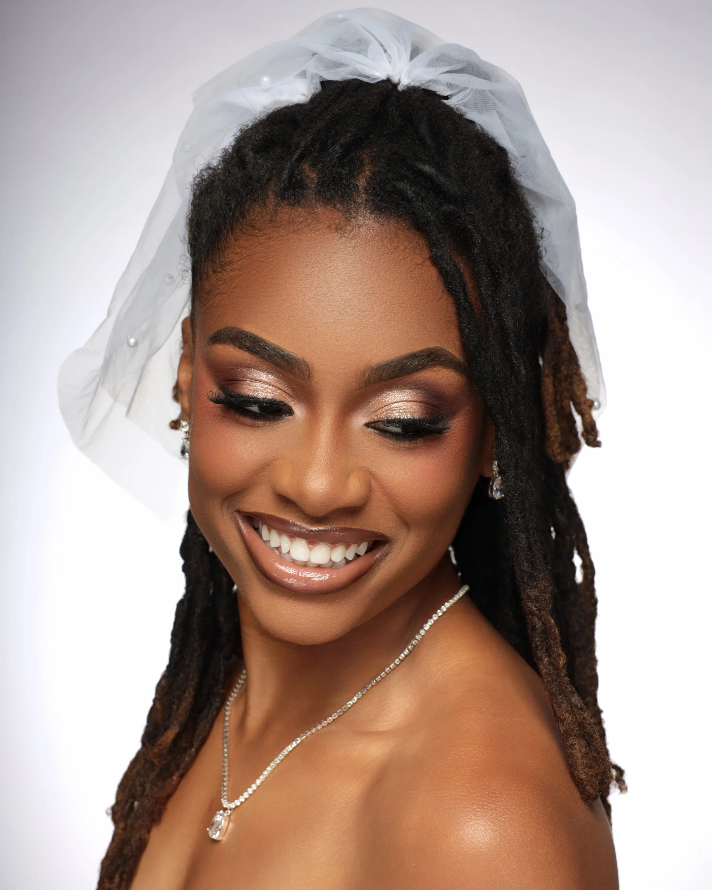 Close-up of a woman with styled dreadlocks, wearing a white tulle veil with pearl embellishments, makeup, earrings, and a necklace, smiling and showing teeth. Bridal makeup