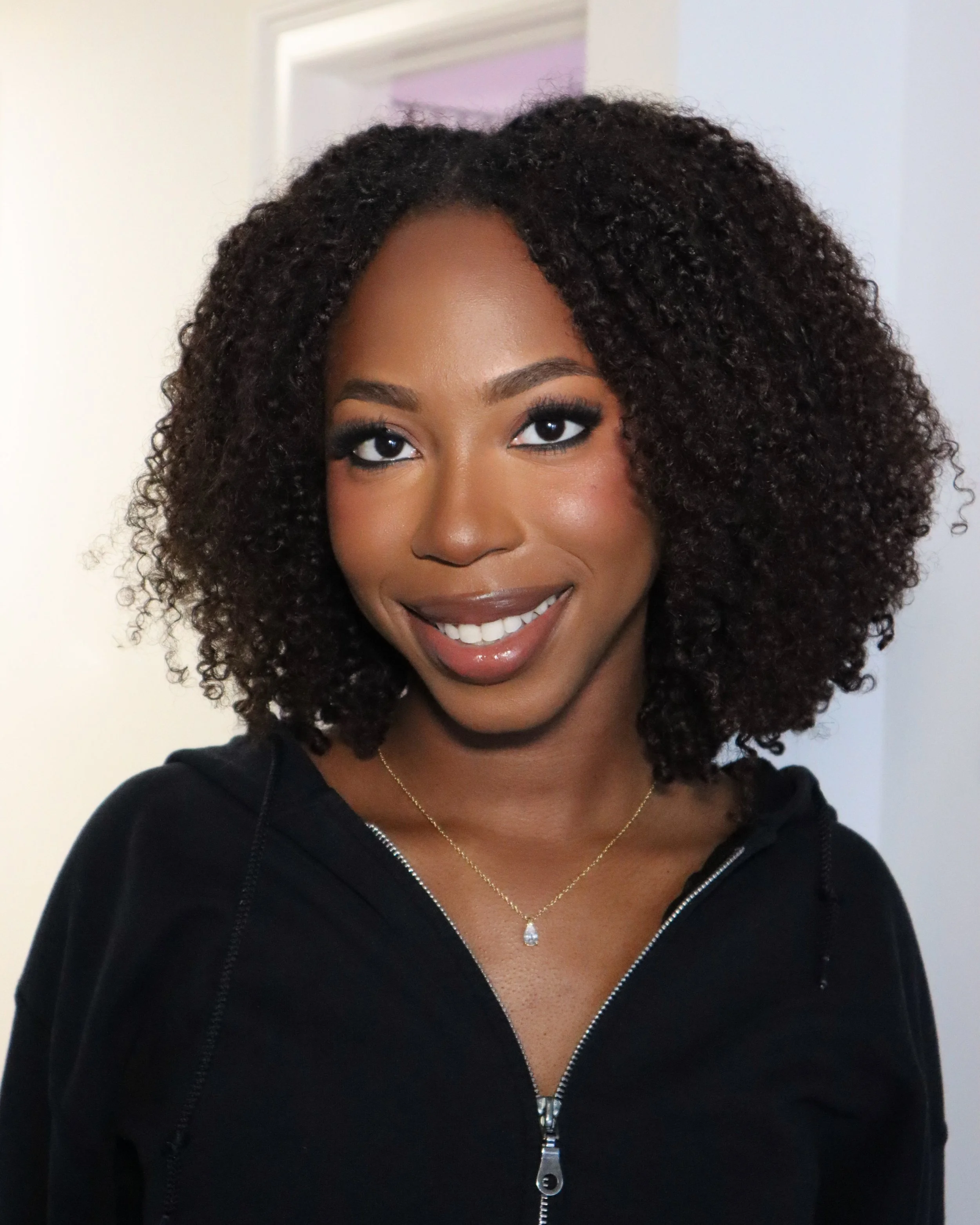 Close-up of a woman with dark skin and curly hair, smiling, wearing a black zip-up jacket, a delicate gold necklace with a small pendant, and makeup including dark eyeshadow and lipstick.