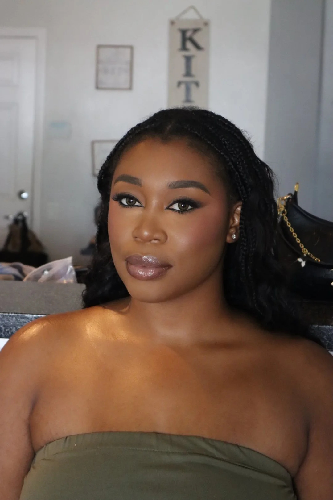 A young woman with dark braided hair and makeup, sitting indoors in front of a white wall with decorative items, wearing a strapless green top.