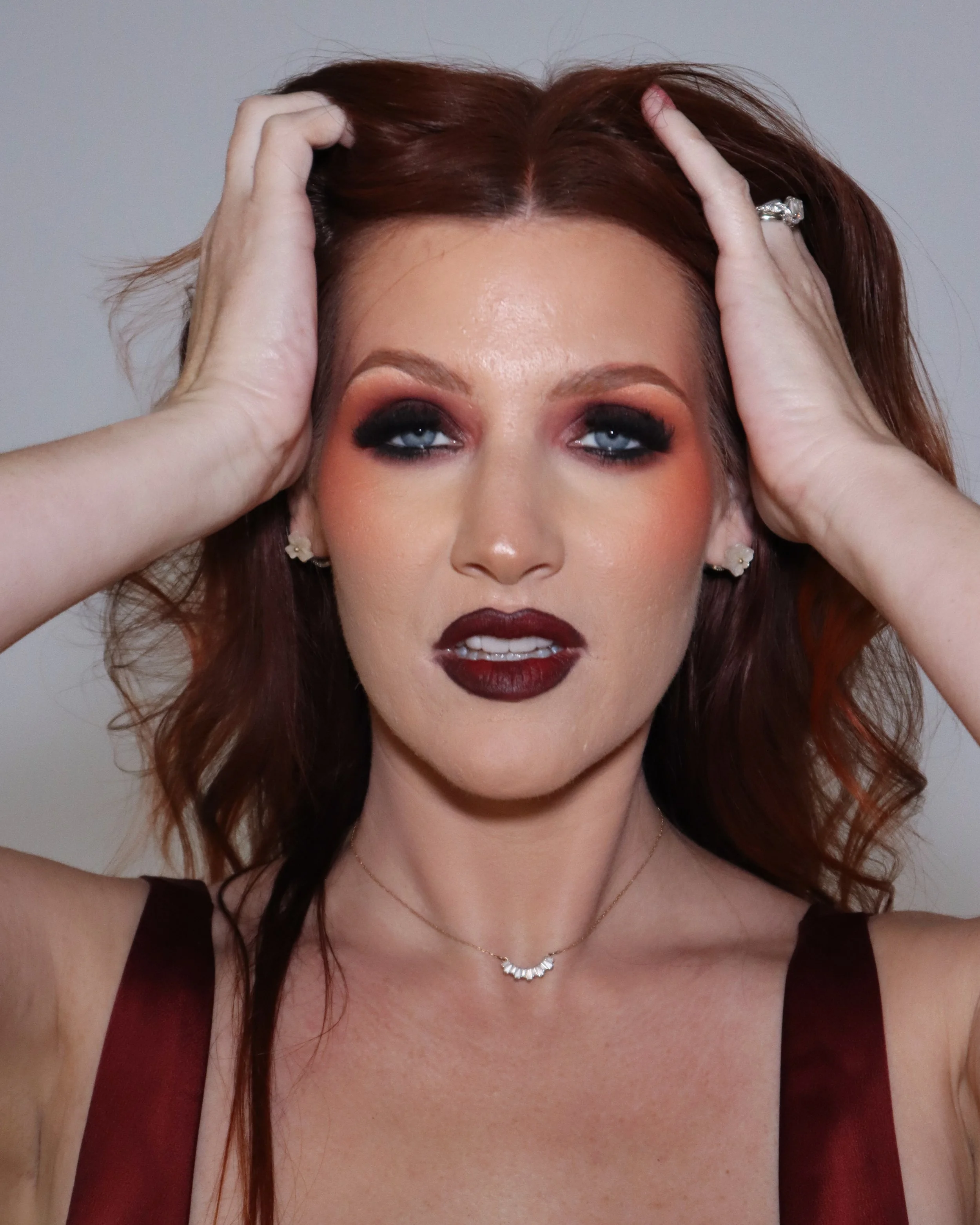 A woman with red hair and blue eyes, wearing dark makeup, a maroon dress, a necklace, and a ring, holding her hair back with both hands.