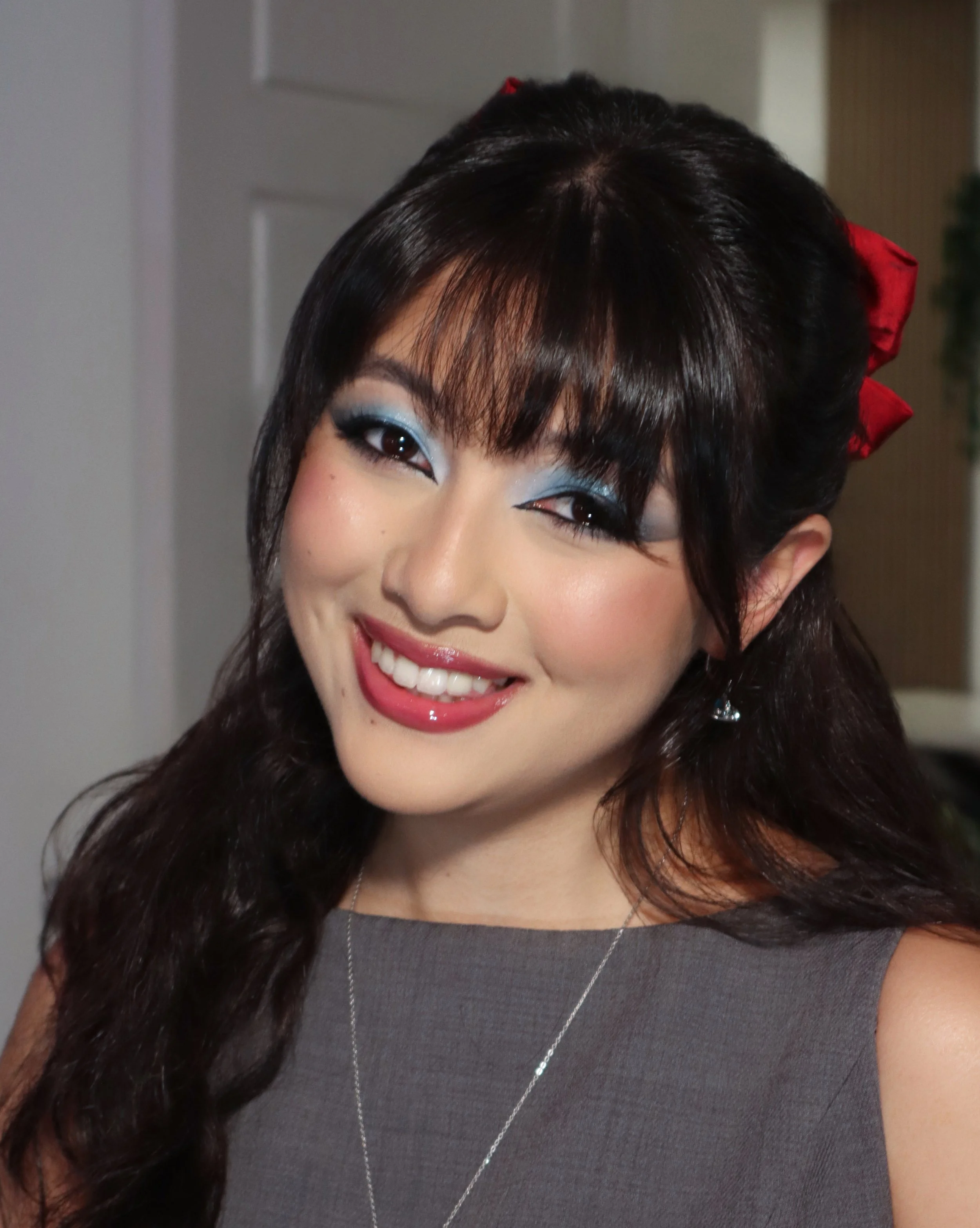 Close-up of a smiling woman with dark hair, wearing blue eyeshadow, a dark gray top, a silver necklace, and a red ribbon in her hair.