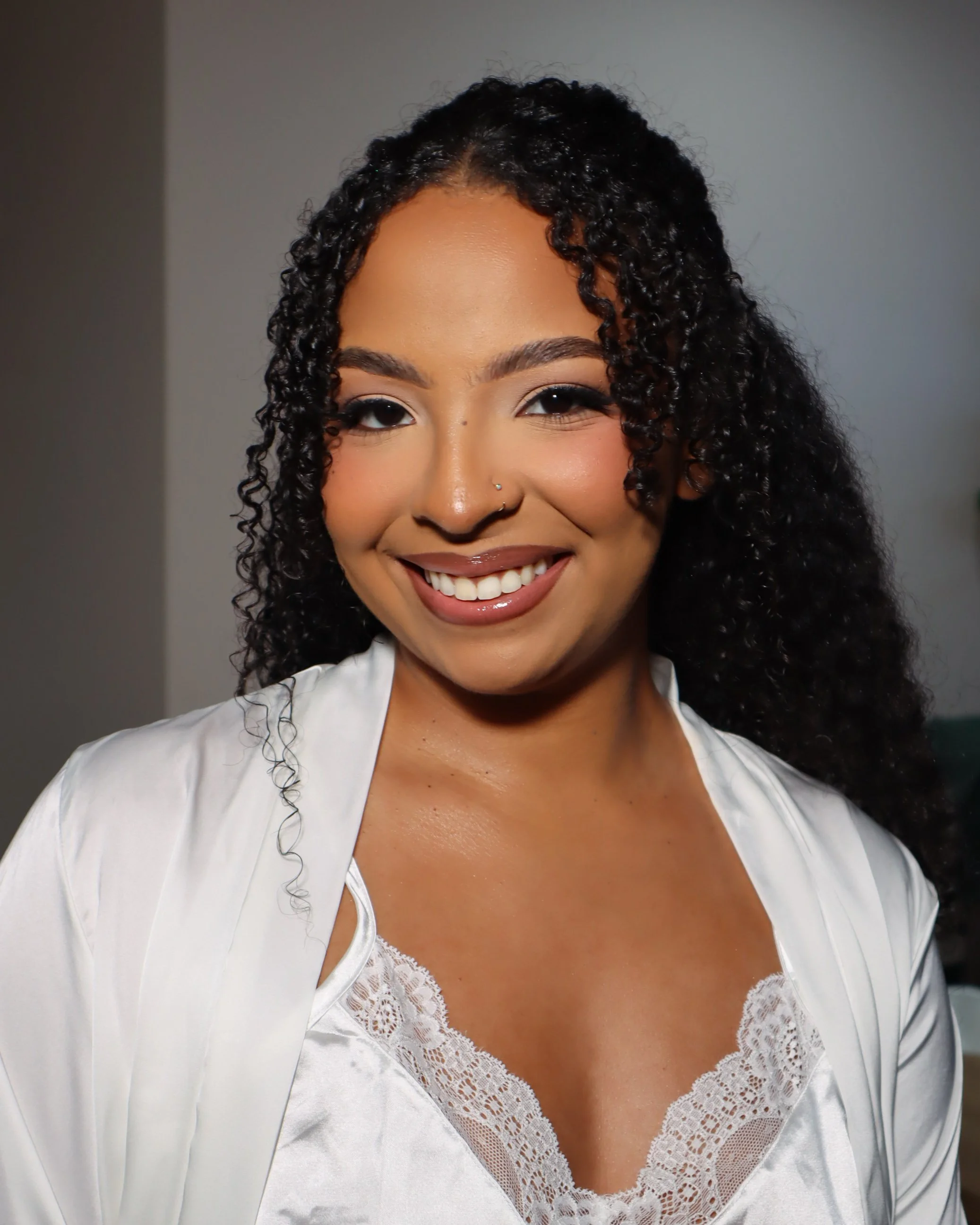 Close-up of a woman's face with curly hair, smiling, wearing a white lace top and a white jacket, with a neutral background.