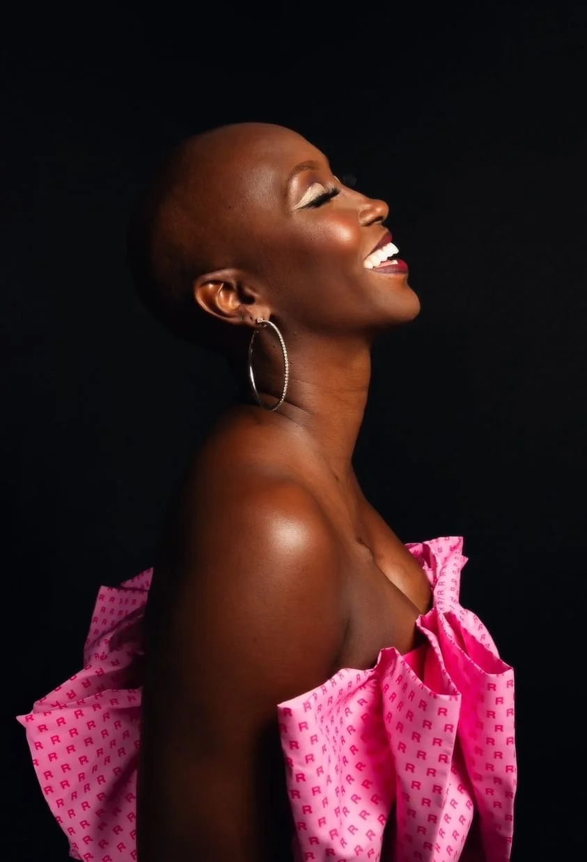 A joyful African American woman with a shaved head, wearing hoop earrings and pink off-shoulder fabric, smiling with her eyes closed against a black background.