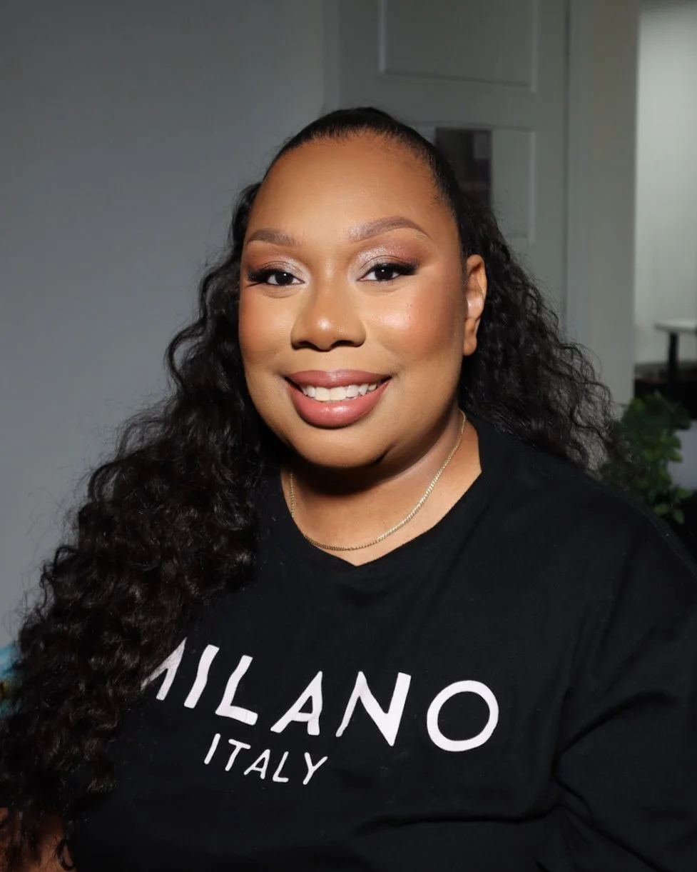 A smiling woman with long, curly black hair, wearing a black T-shirt with the words 'MILANO ITALY' on it, standing indoors.