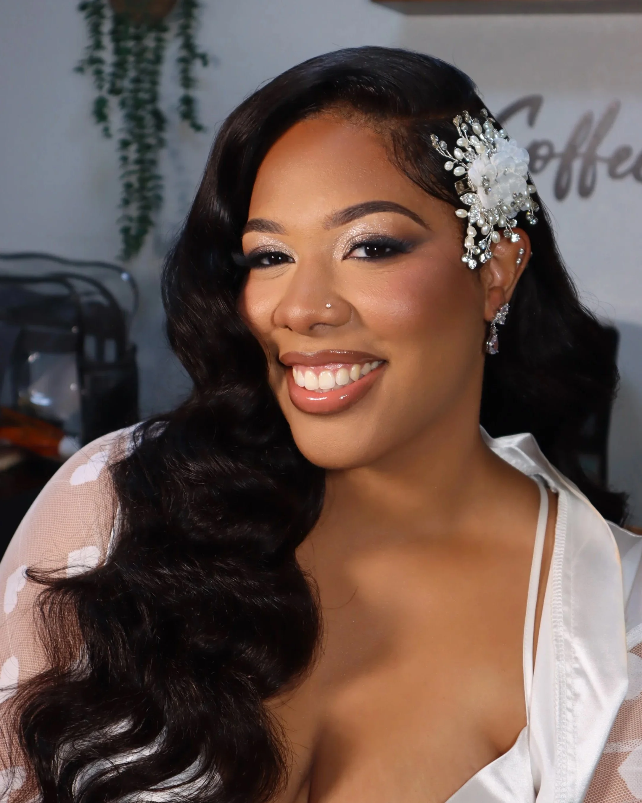A woman with long, dark, curled hair styled to one side, wearing a decorative pearl and crystal hair accessory, sparkling eye makeup, and a nose piercing, smiling while sitting indoors. Bridal makeup