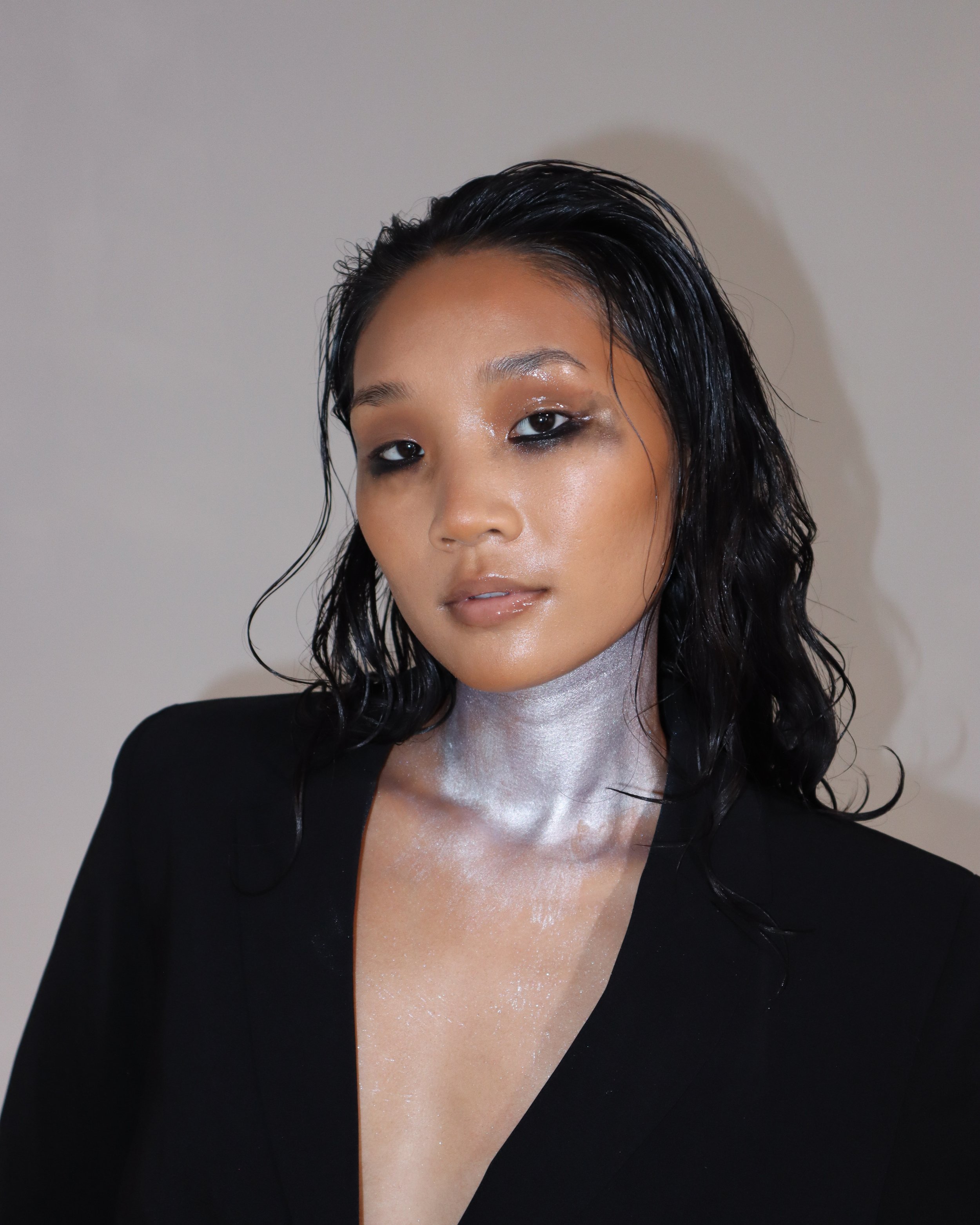Close-up of a woman with wet black hair, wearing smudged eye makeup and a black top, against a neutral background.