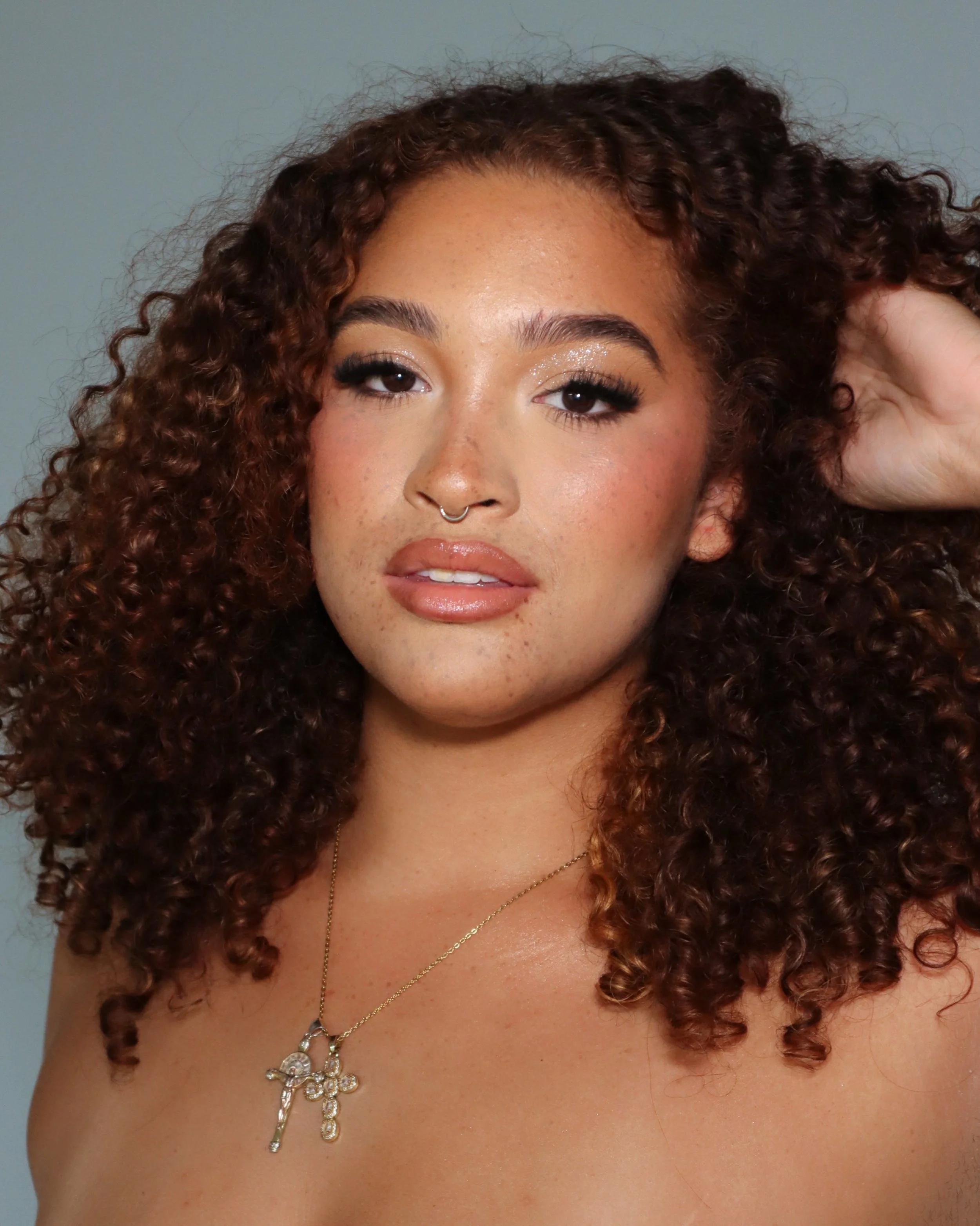Close-up of a woman with long, curly, reddish-brown hair, wearing makeup with glittery eyeshadow, glossy nude lipstick, a septum piercing, and layered necklaces, against a plain background.