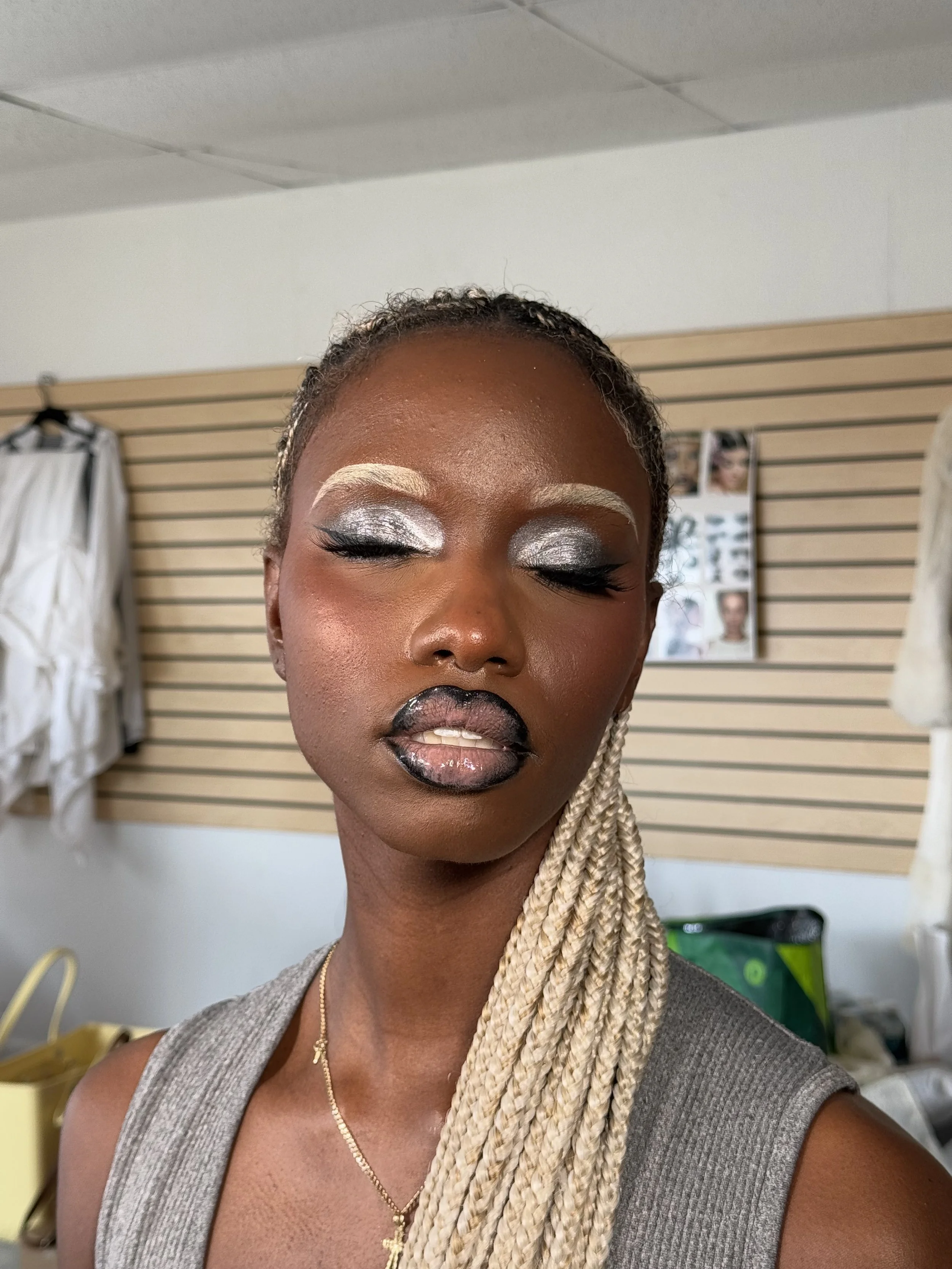 A woman with braided blonde hair is wearing silver and black makeup, including silver eyeshadow, black eyeliner, and black lipstick, with a pearl necklace and gray sleeveless top, in front of a slatwall with posters in the background.