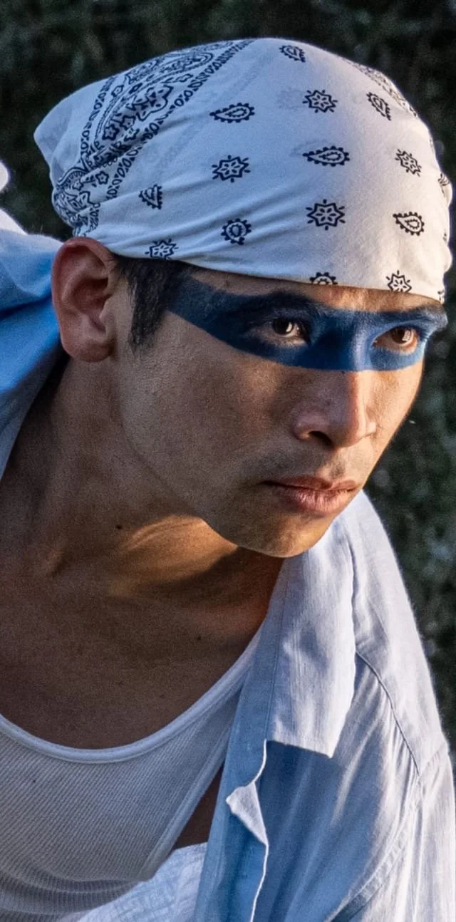 A man with black face paint and a white bandana with black paisley pattern, wearing a white shirt, outdoors at sunset.
