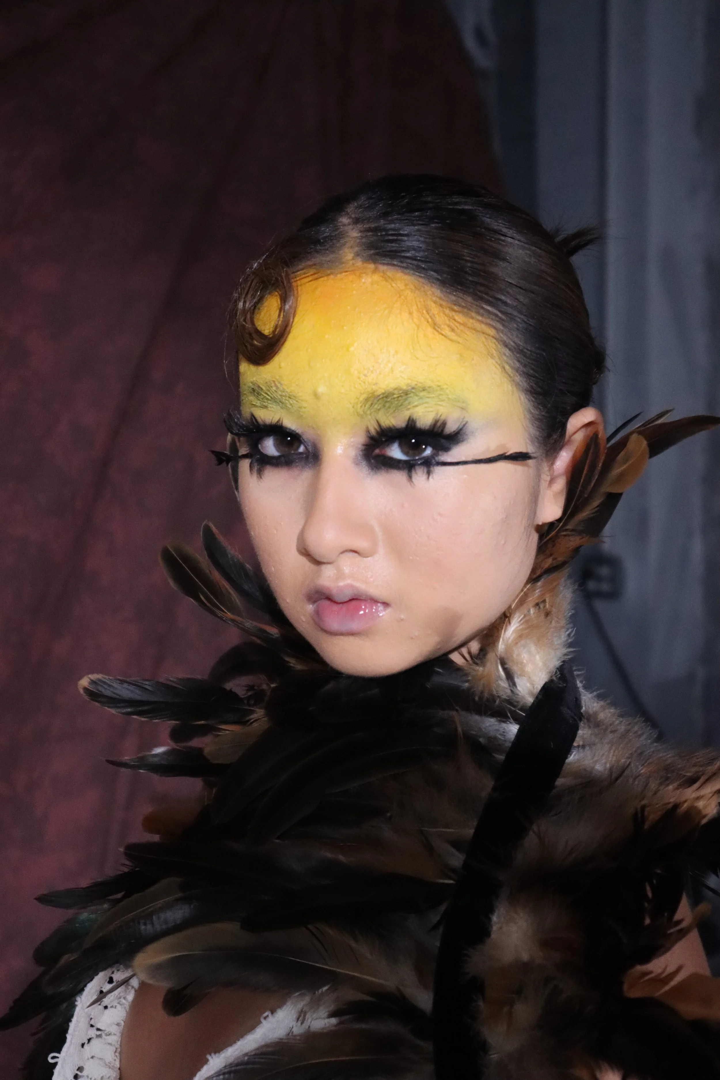 Close-up of a woman with elaborate makeup featuring yellow face paint, dramatic black eyeliner, and long eyelashes, wearing a costume with feathers and fur, against a dark background.