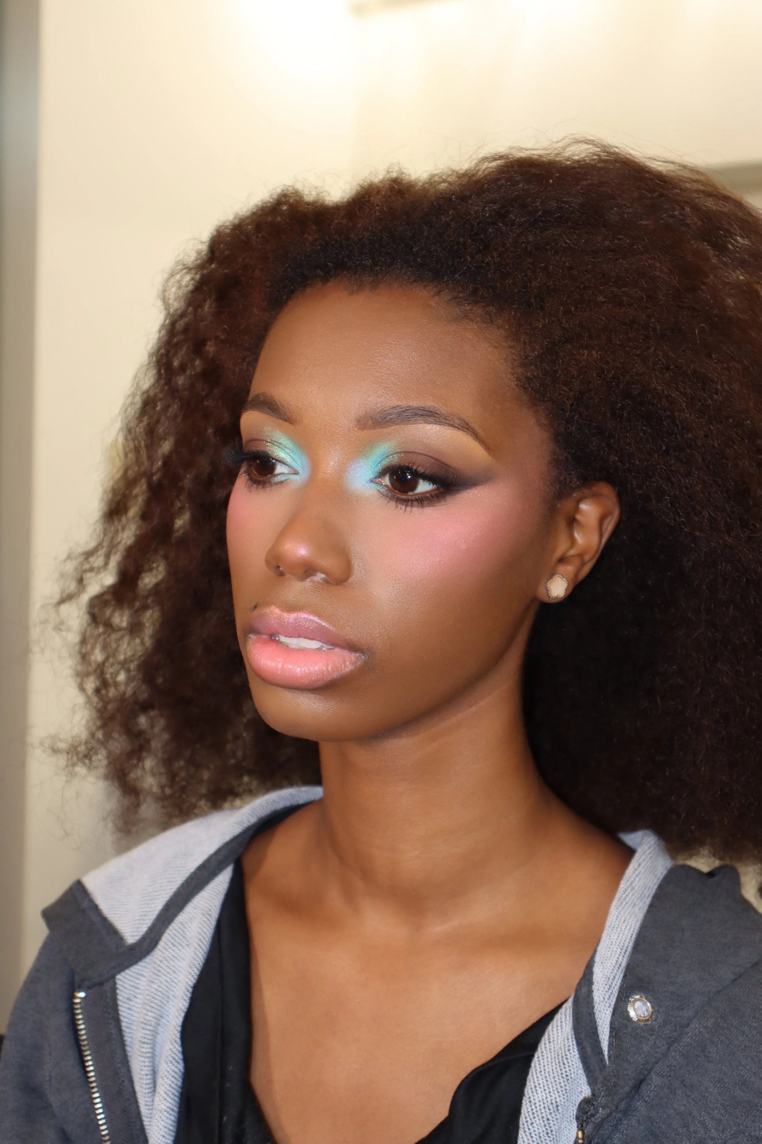 Close-up of a woman with natural curly hair, wearing colorful makeup including blue eyeshadow and pink blush, and a casual hoodie.