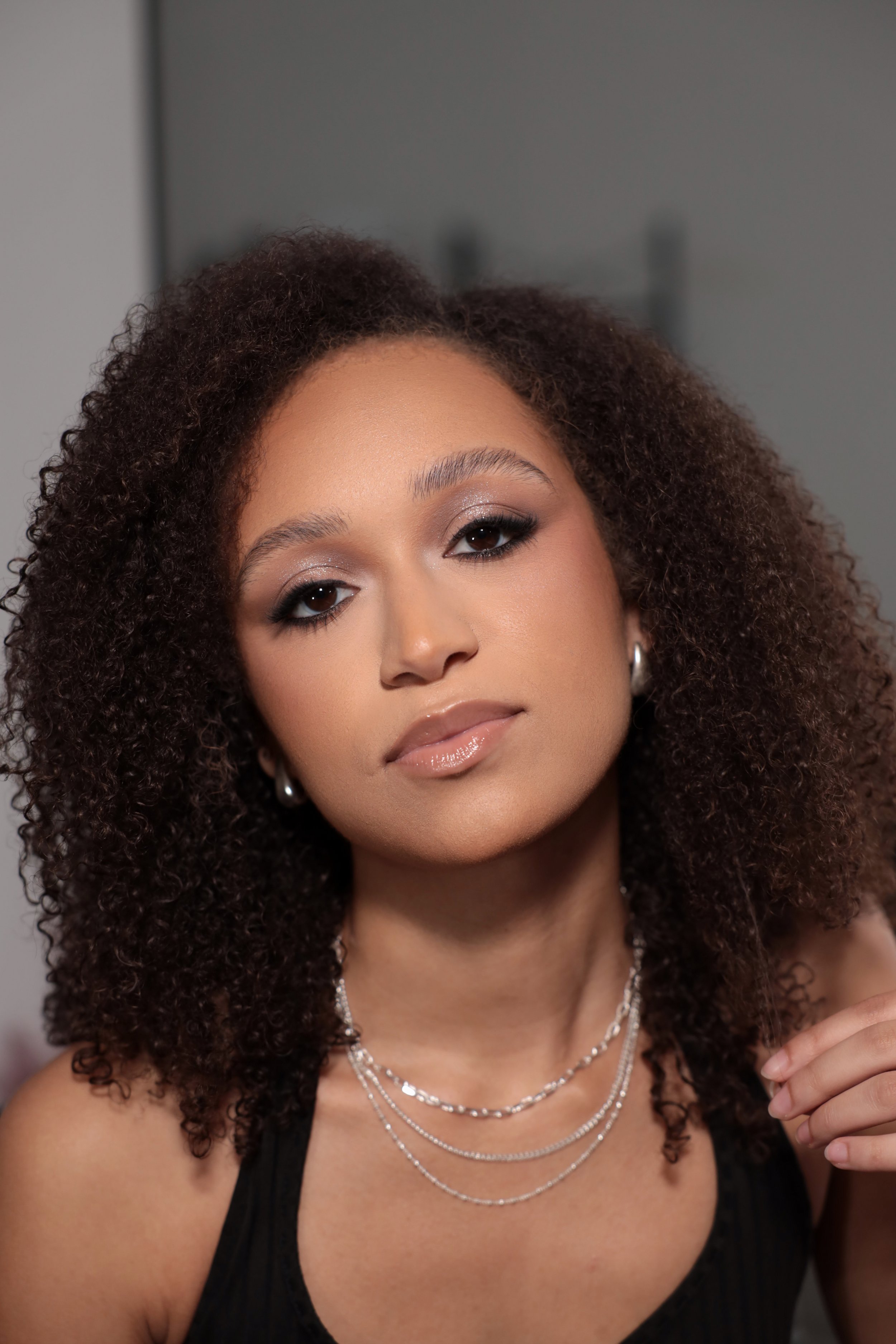 Close-up of a woman with curly hair, wearing makeup, silver jewelry, and a black top, posing indoors.