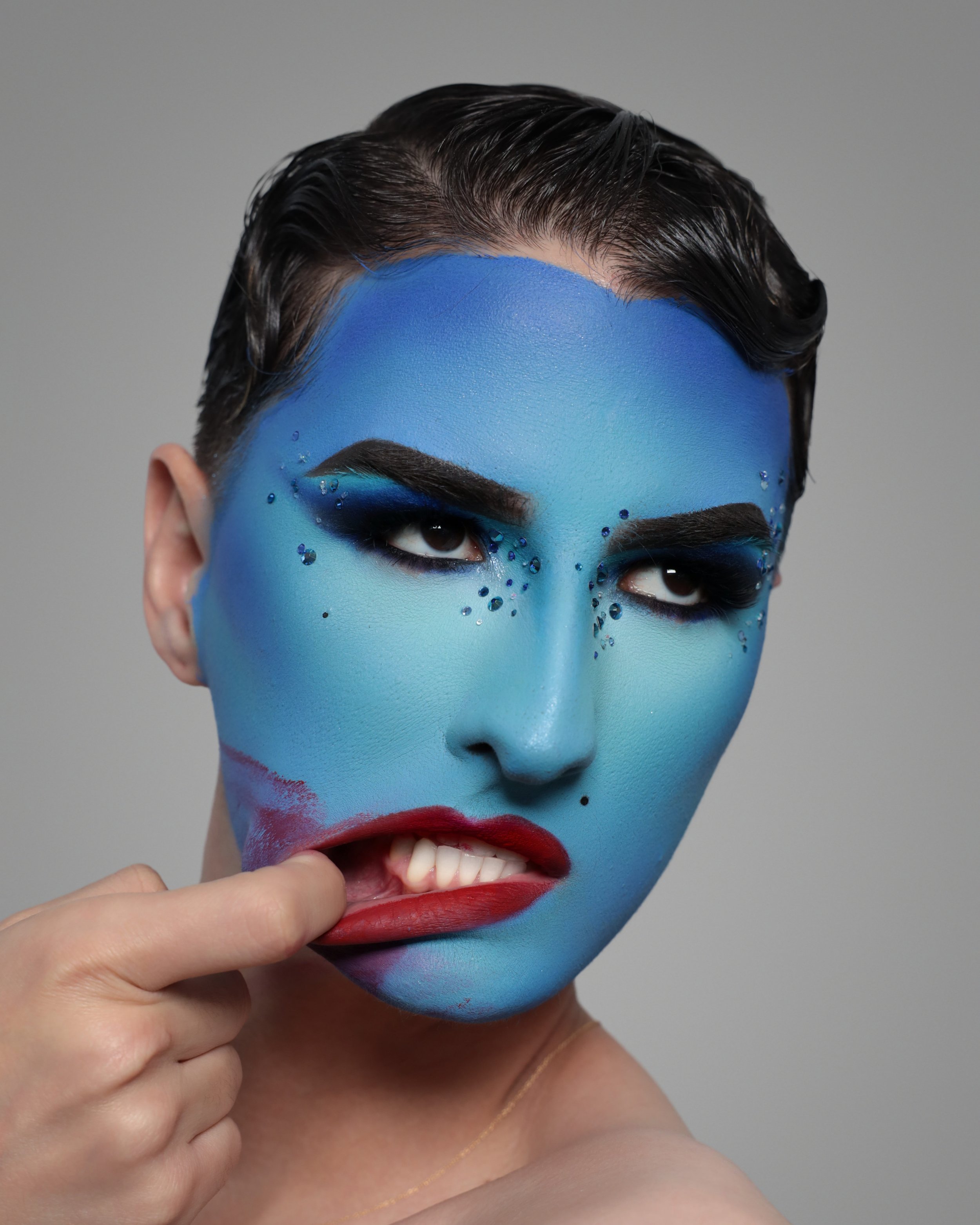 Close-up of a person's face with artistic makeup. Their face is painted blue with accents of darker blue, with rhinestones and sparkles, and bold red lipstick. They are touching their chin with their hand, and have dark, slicked-back hair, and facial