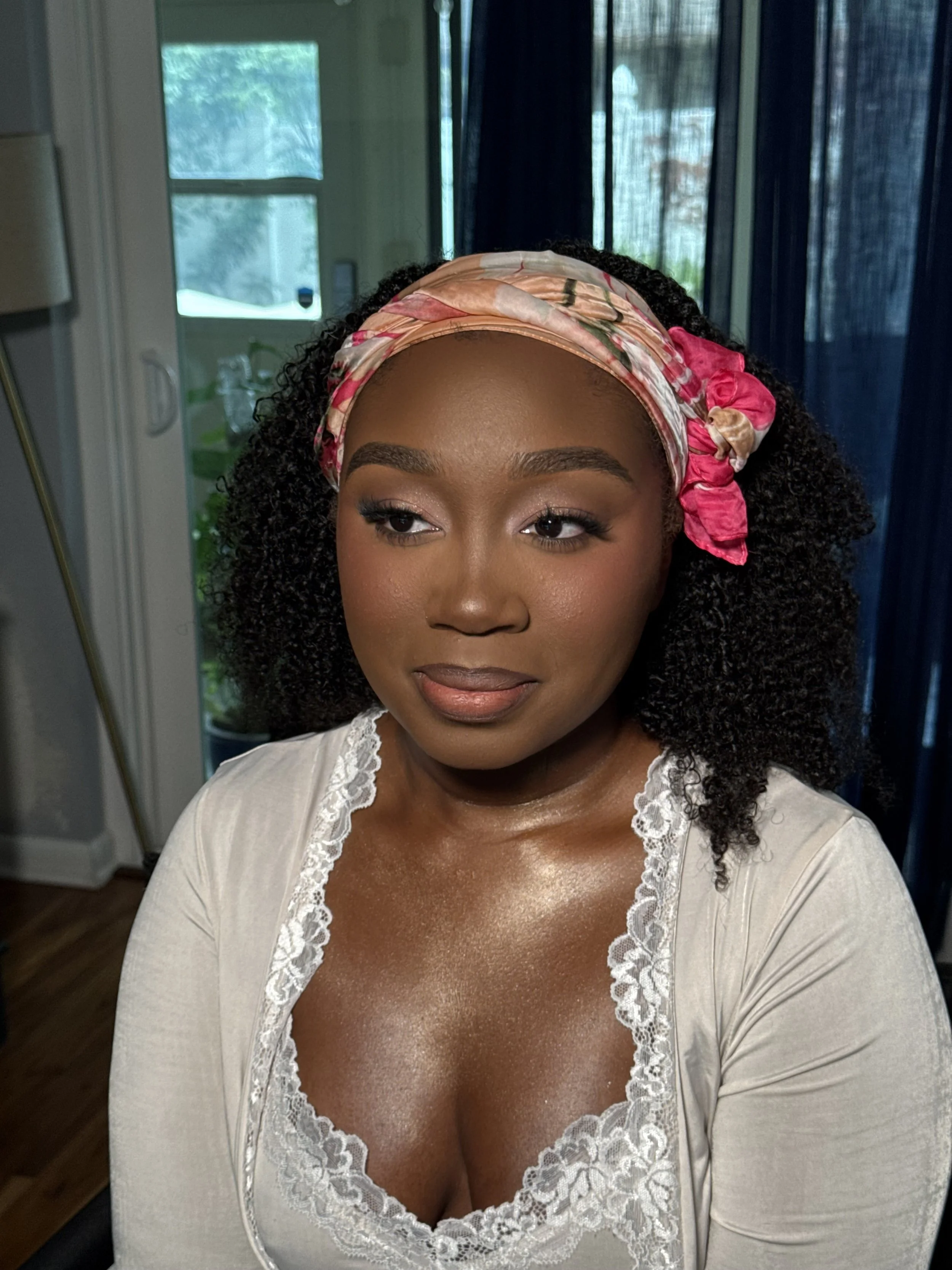A woman with dark curly hair wearing a pink satin headscarf with floral patterns and a beige top with lace details, posing indoors near a glass door with a green outdoor view.