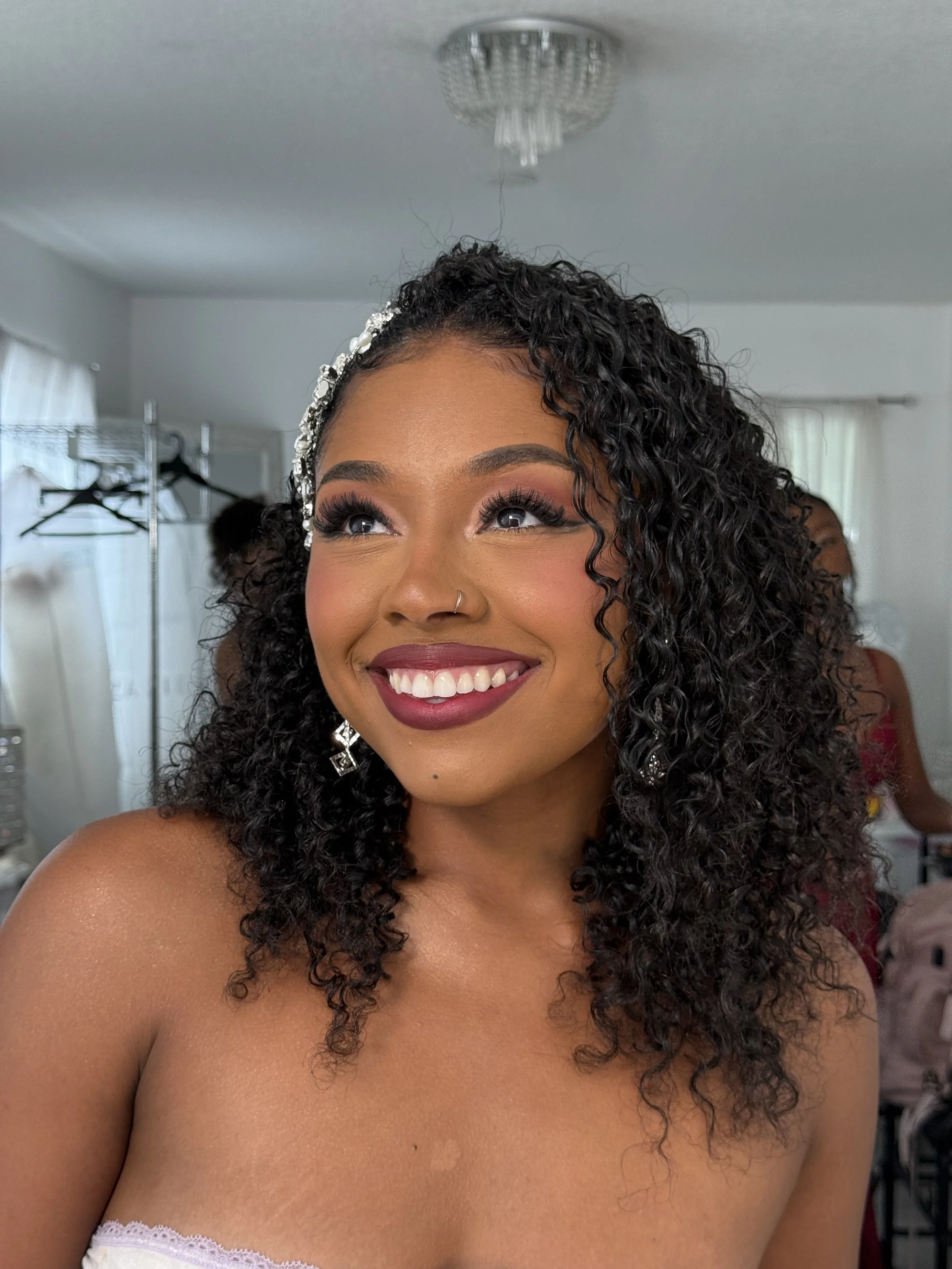 Close-up of a woman with curly hair, wearing makeup, earrings, and a pearl hair accessory, smiling with visible teeth, in a room with a ceiling light and closet in the background.