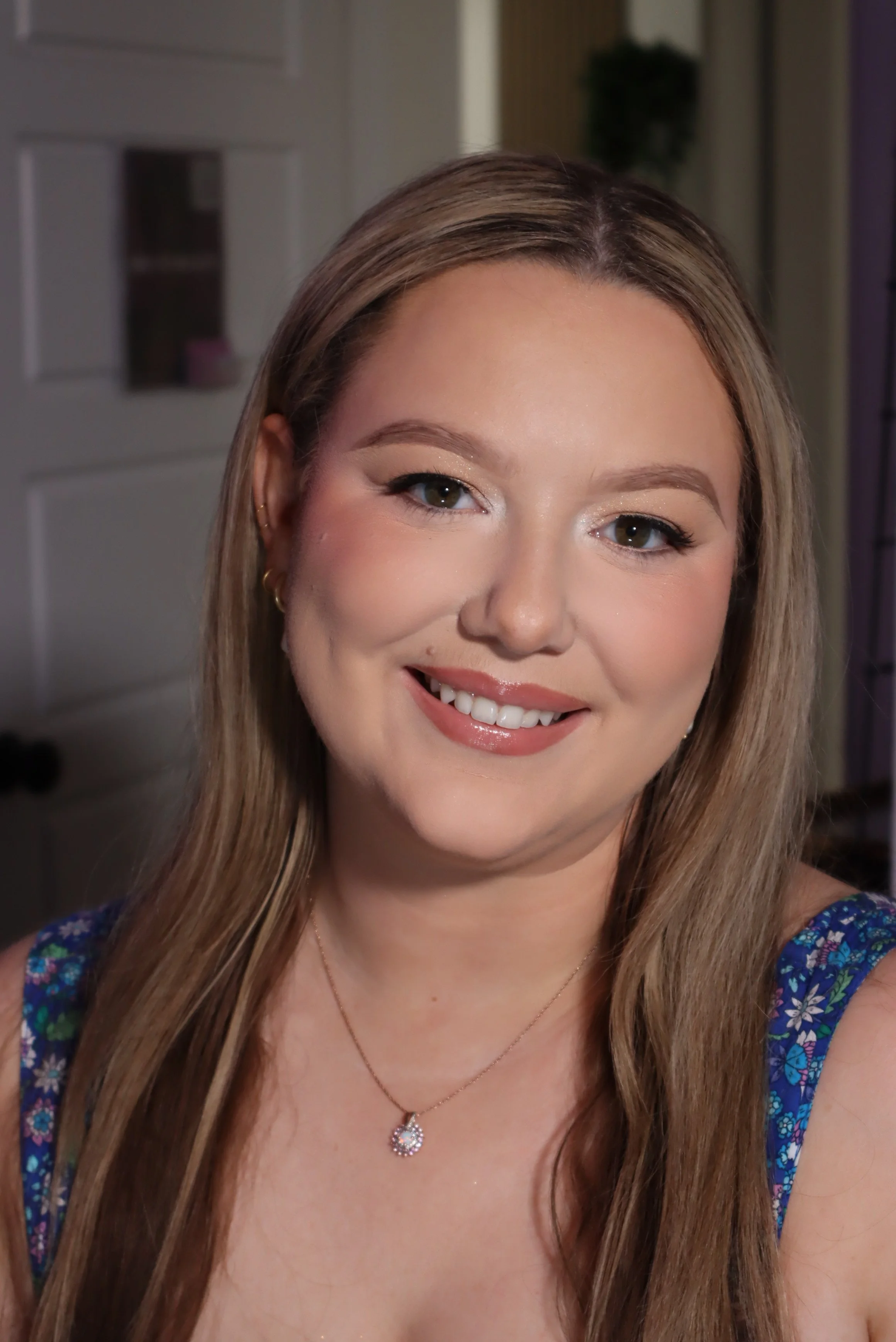 Close-up of a smiling woman with long, light brown hair, wearing makeup, a necklace with a teardrop-shaped pendant, and a blue floral dress, standing indoors.