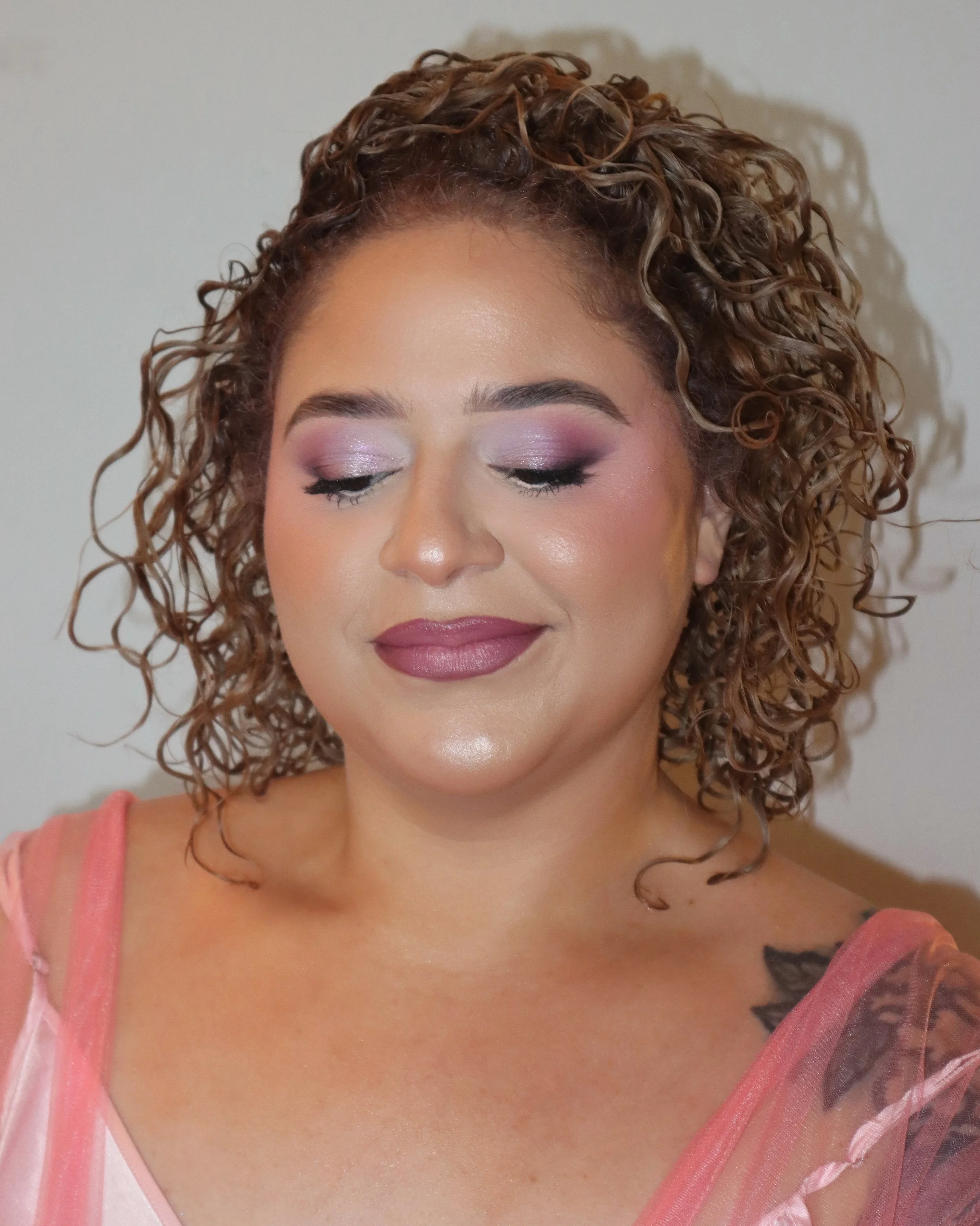 Close-up of a woman with curly hair, wearing pink makeup and a pink sheer top, with her eyes closed.