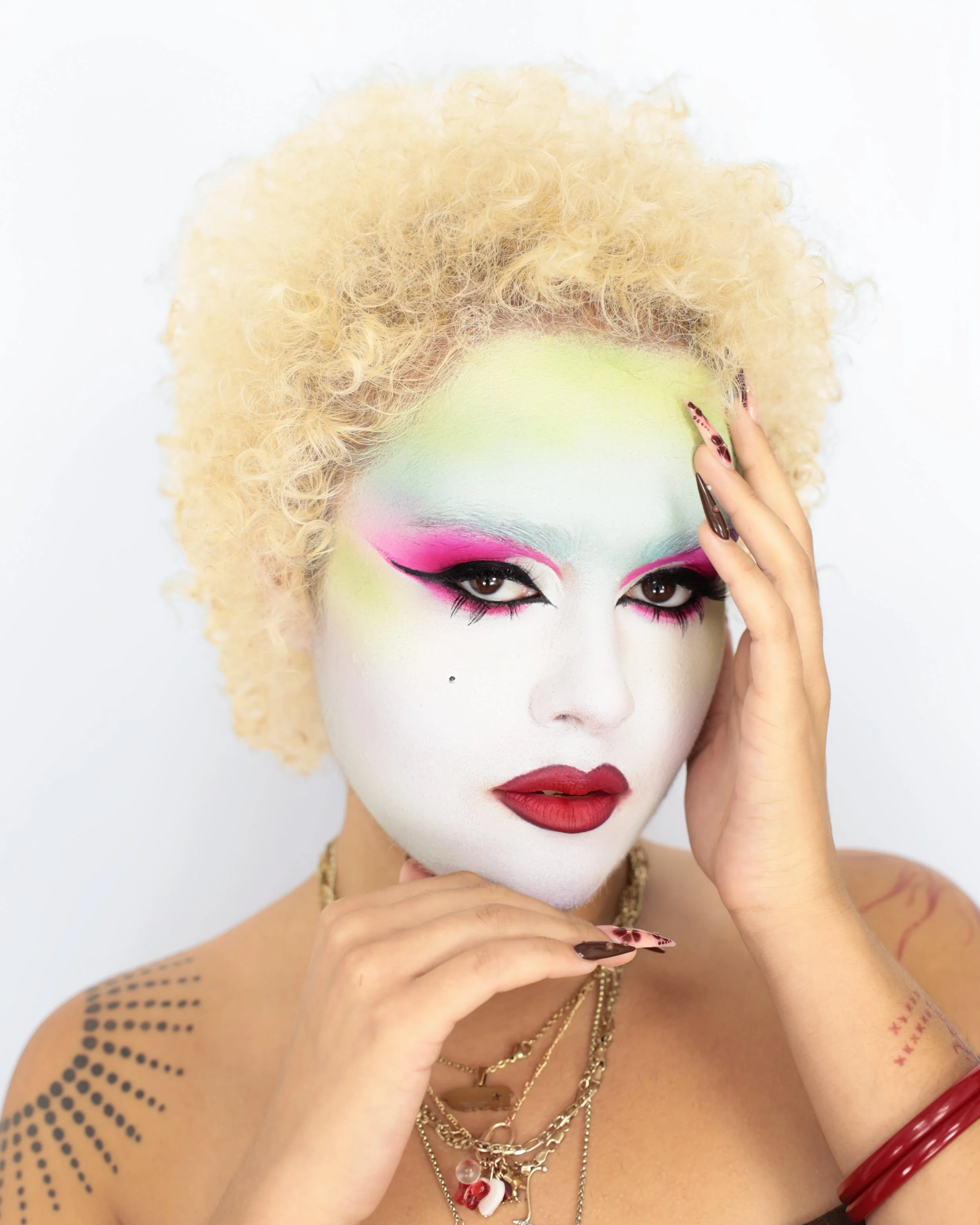 A person with pale skin and bright, colorful makeup, wearing multiple layered gold necklaces, red and black nail polish, and a red bangle. They have platinum blonde curly hair and are touching their face with one hand, with a white background.
