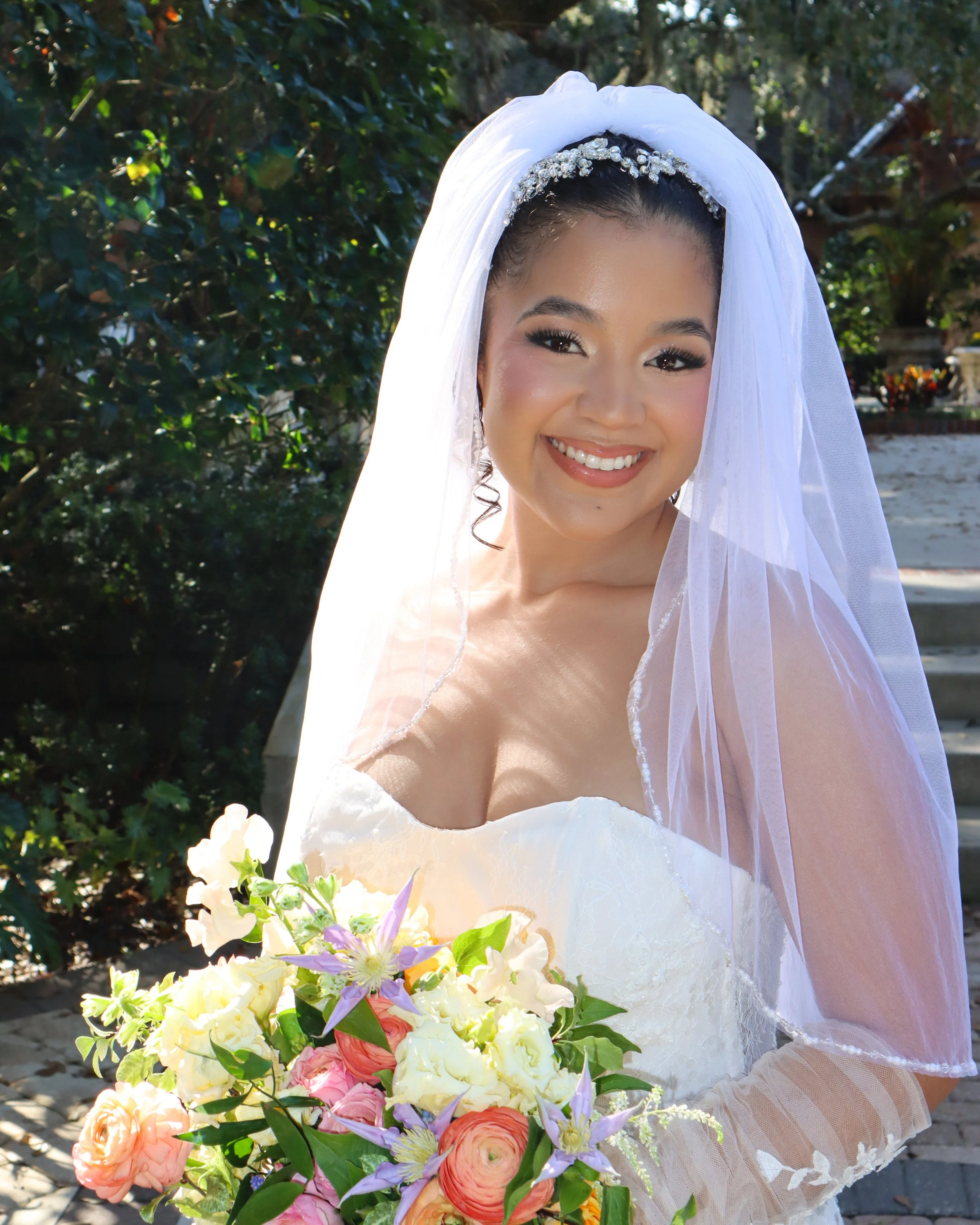 Young woman dressed as a bride, wearing a white wedding dress with lace and an embellished headband, smiling outdoors. She holds a large bouquet of mixed flowers, including pink roses, purple tulips, and white and yellow blooms, with trees and sunlig