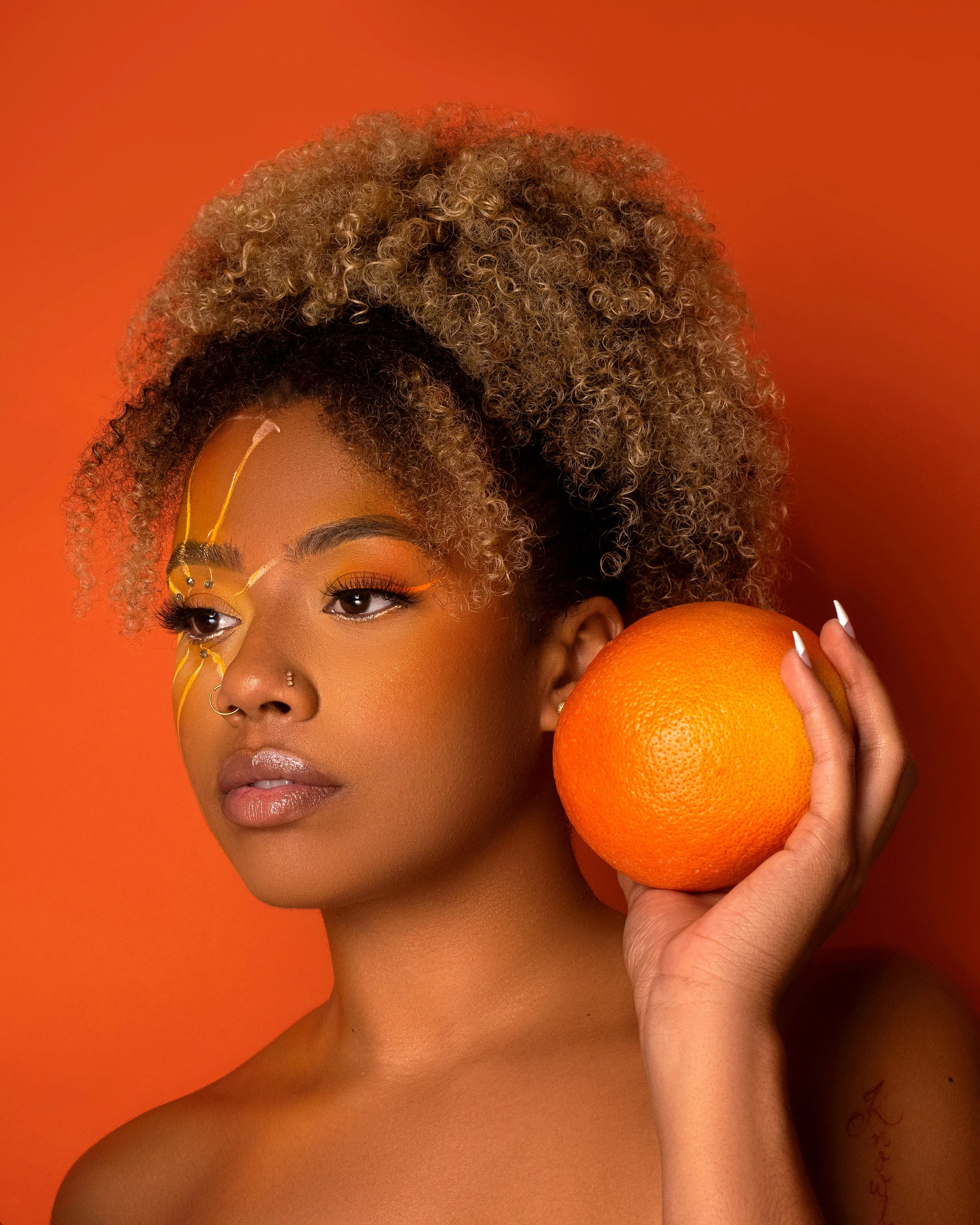 Close-up of a woman with curly blonde hair holding an orange fruit next to her face against an orange background.