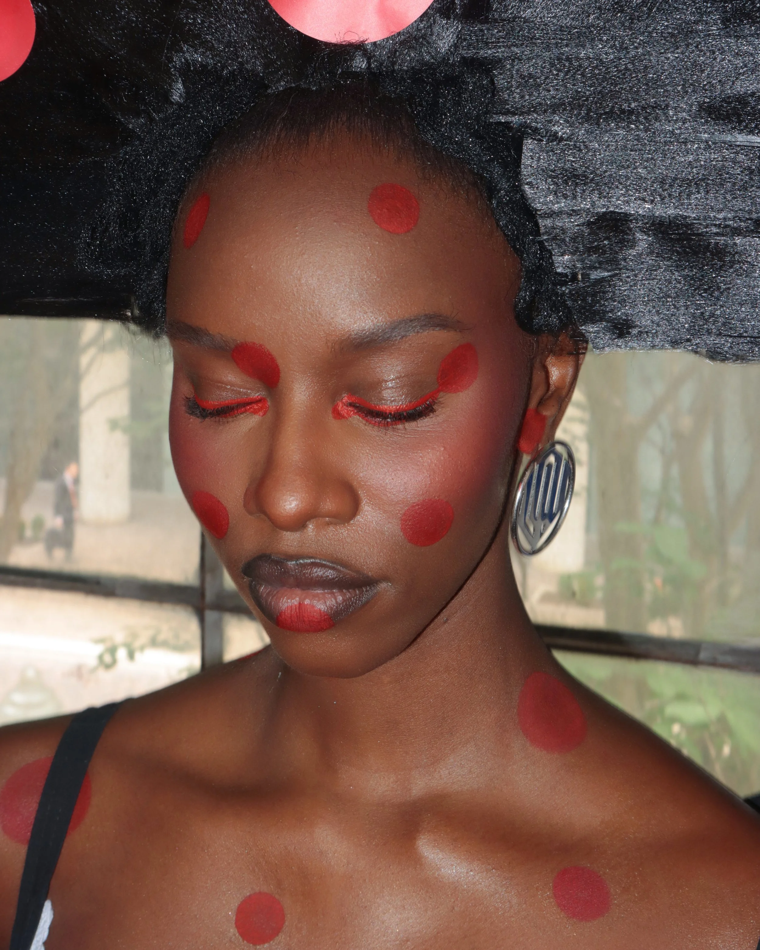 A woman with dark skin and short hair has red circles painted on her face, neck, and shoulders. She is wearing earrings and has her eyes closed.