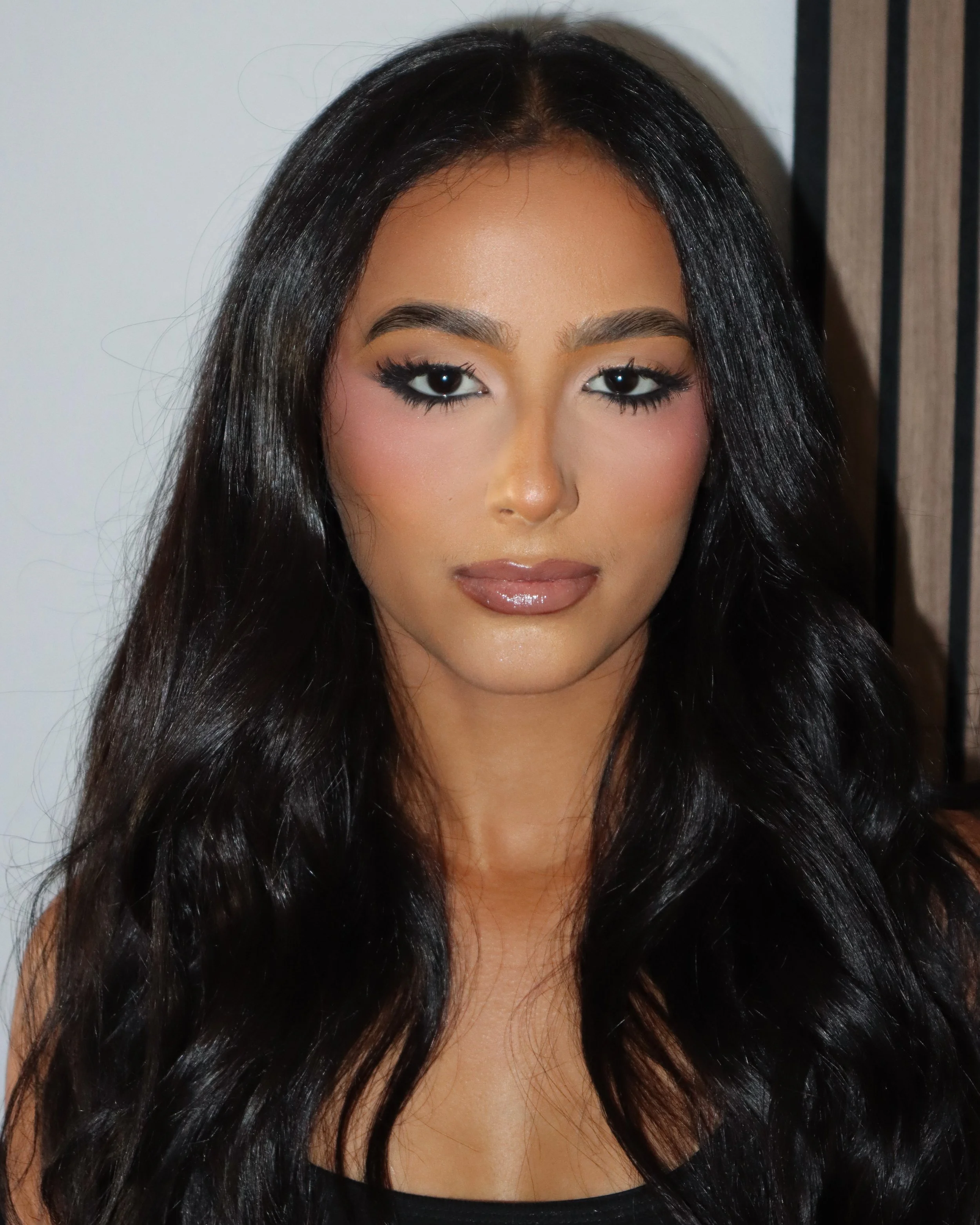 A woman with long, shiny black hair, makeup including dark eyeliner and lipstick, wearing a black top, standing against a plain background with a curtain on the side.