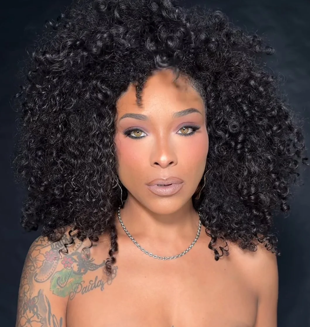 A woman with dark, curly hair, wearing a silver necklace and earrings, staring directly at the camera with a neutral expression.