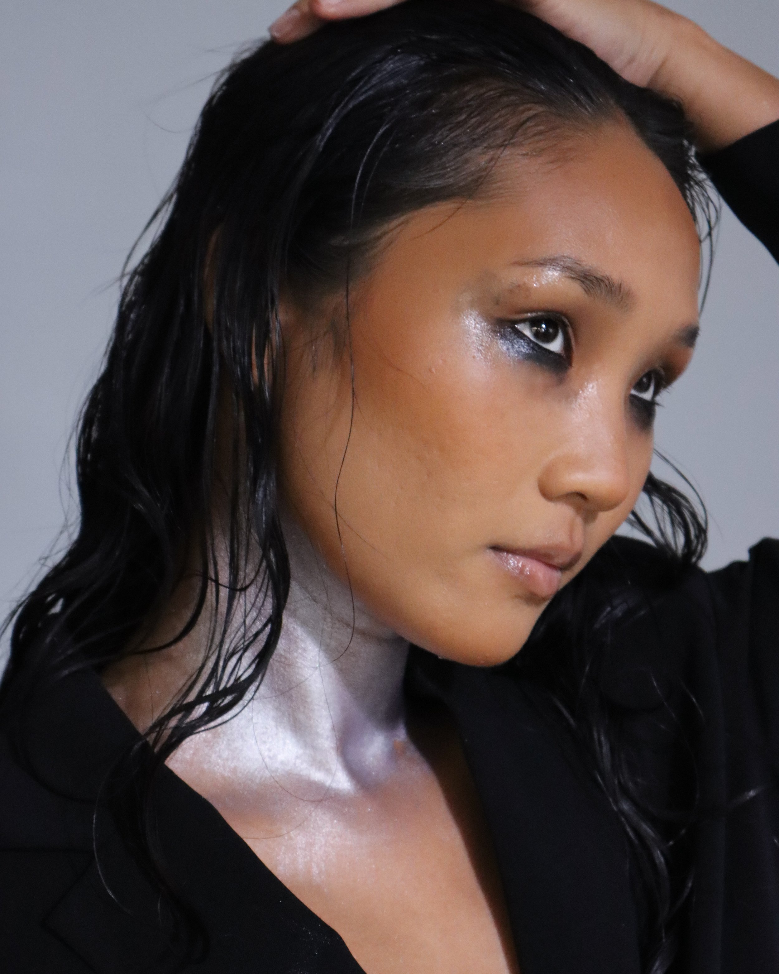 Close-up of a person's face with wet, black hair styled to the side, skin with a natural glow, wearing eye makeup, and a black top.